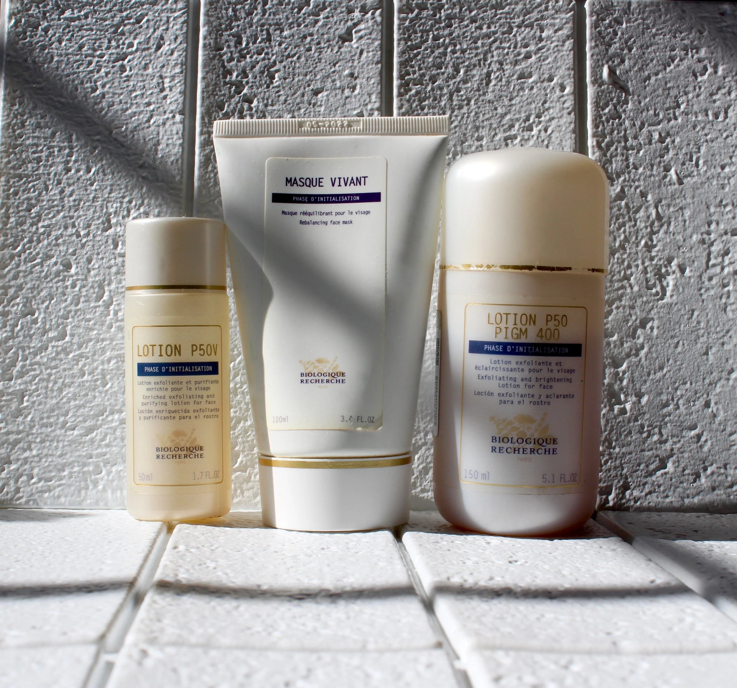 Four white skincare or cosmetic bottles placed on a tiled surface with sunlight casting shadows, against a textured white wall background.