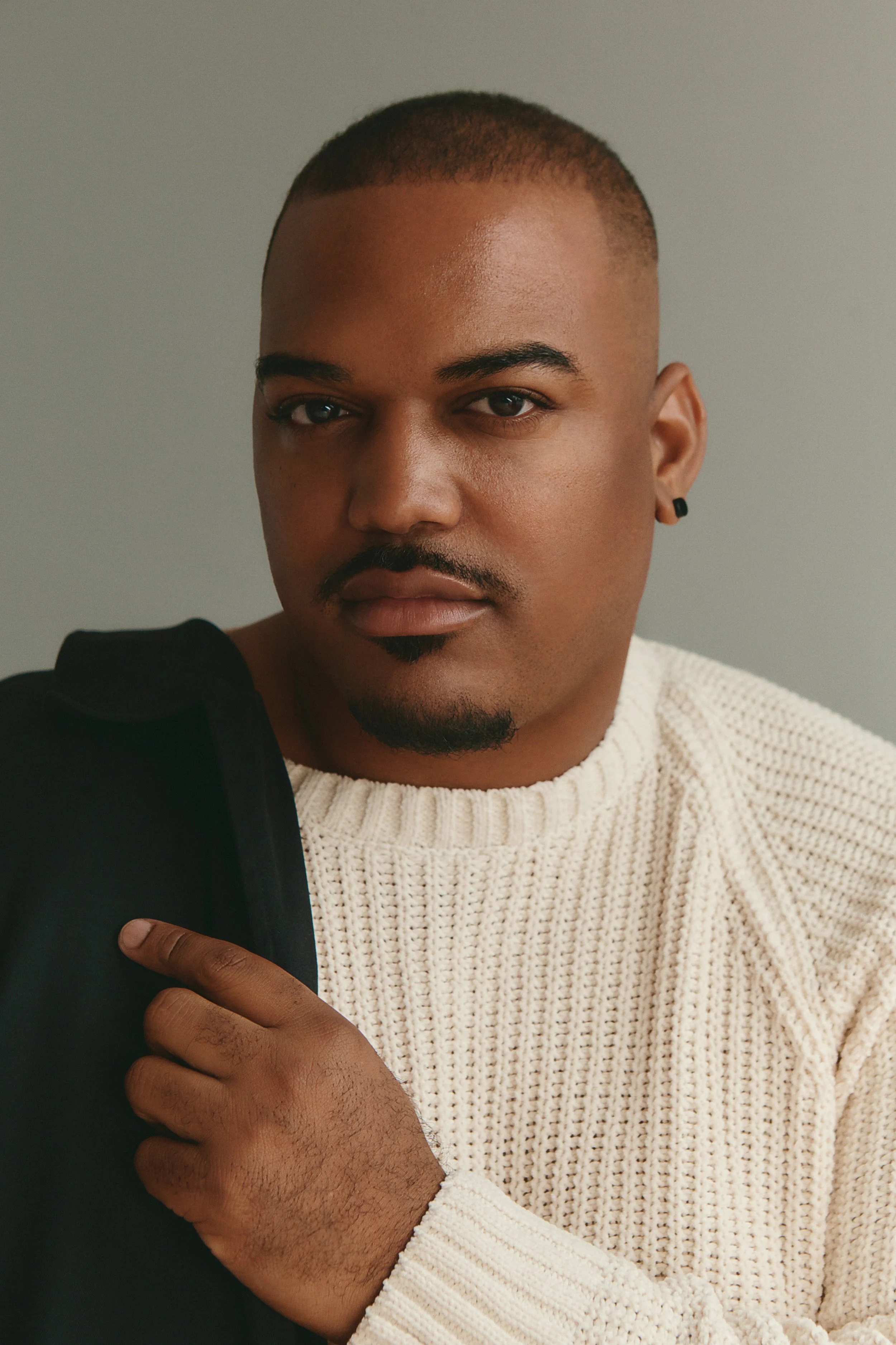 Close-up of a young man with short hair, light brown skin, a well-groomed beard, wearing a cream-colored knit sweater and black earrings, holding a black object, against a plain, light-colored background.