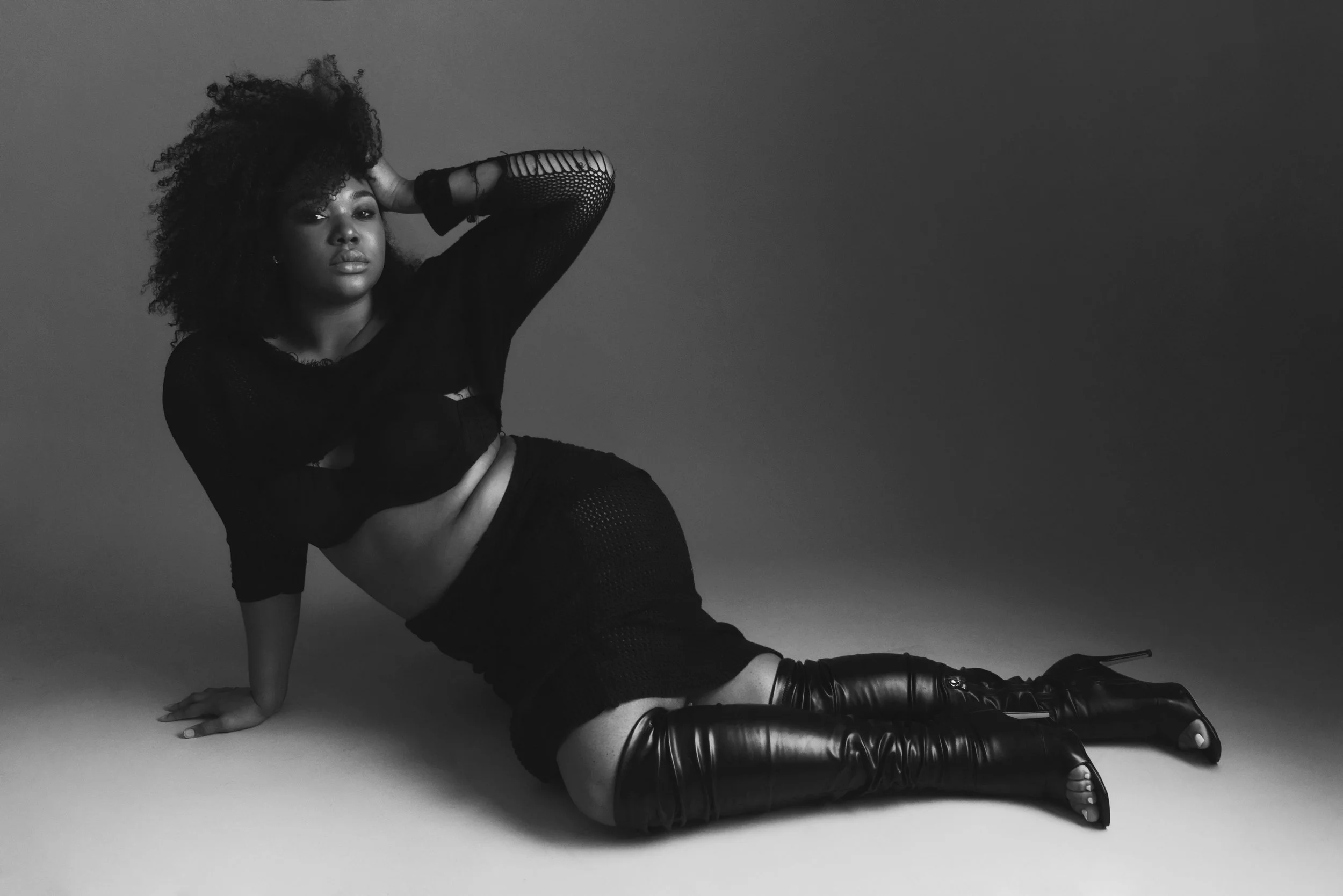 A black and white photo of a woman with curly hair, wearing a black mesh top and shorts, posed on the floor against a plain background.