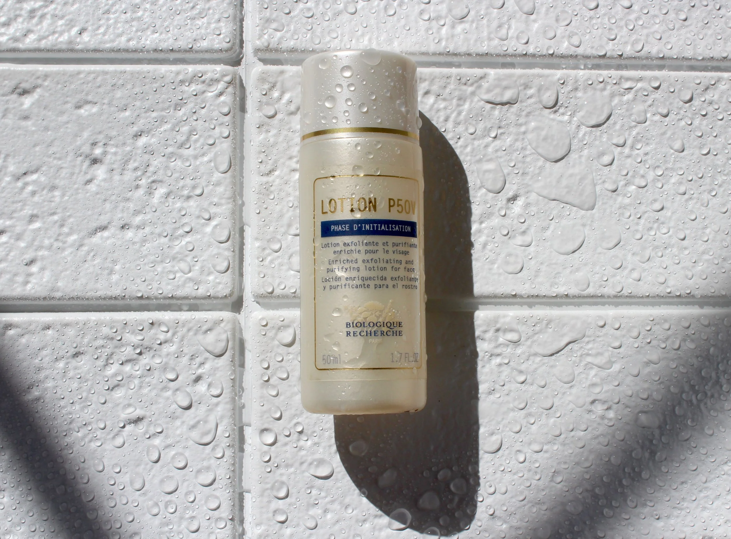 A white bottle of exfoliating and purifying face lotion with a gold band on a white tiled wall covered in water droplets.