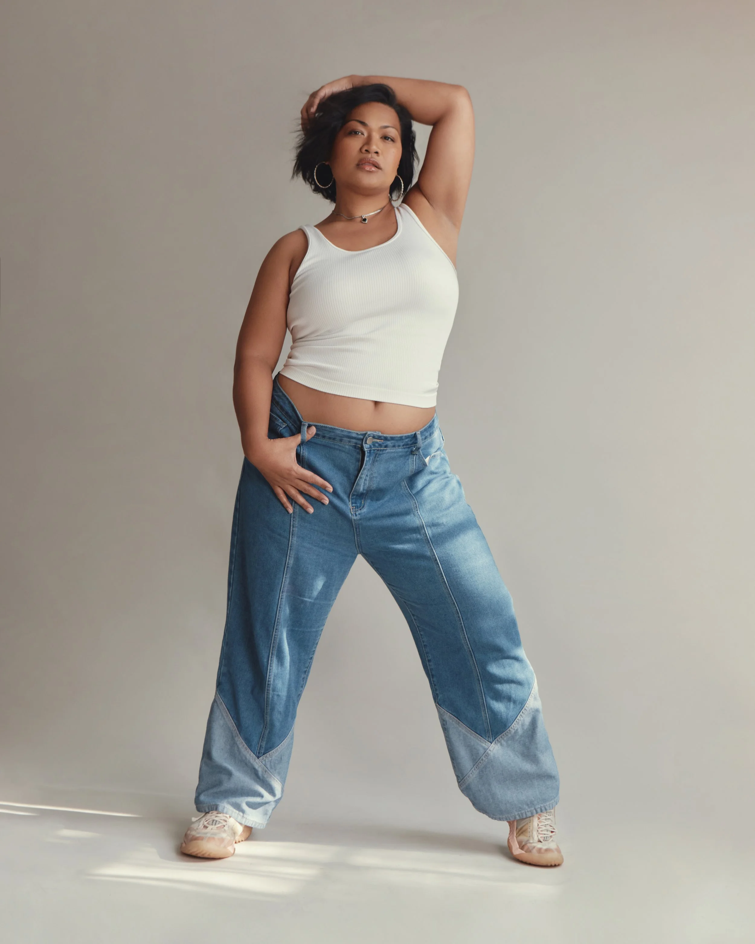 A woman with short black hair wearing a white sleeveless crop top, blue jeans, and pink sneakers, striking a pose with her right hand behind her head and left hand on her hip against a plain gray background.