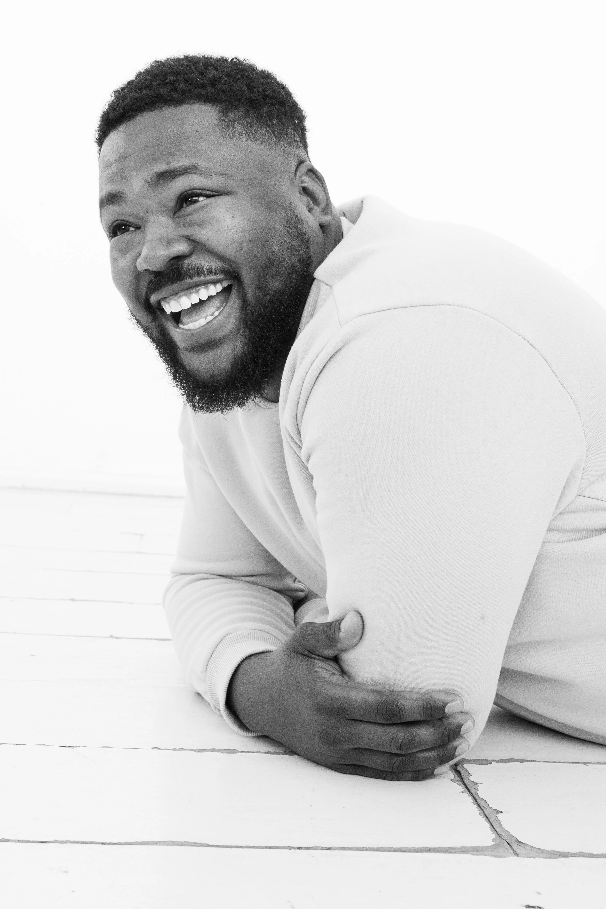 A smiling man with a beard and short hair, wearing a light-colored hoodie, leaning on a white wooden surface, with his arms crossed and laughing.