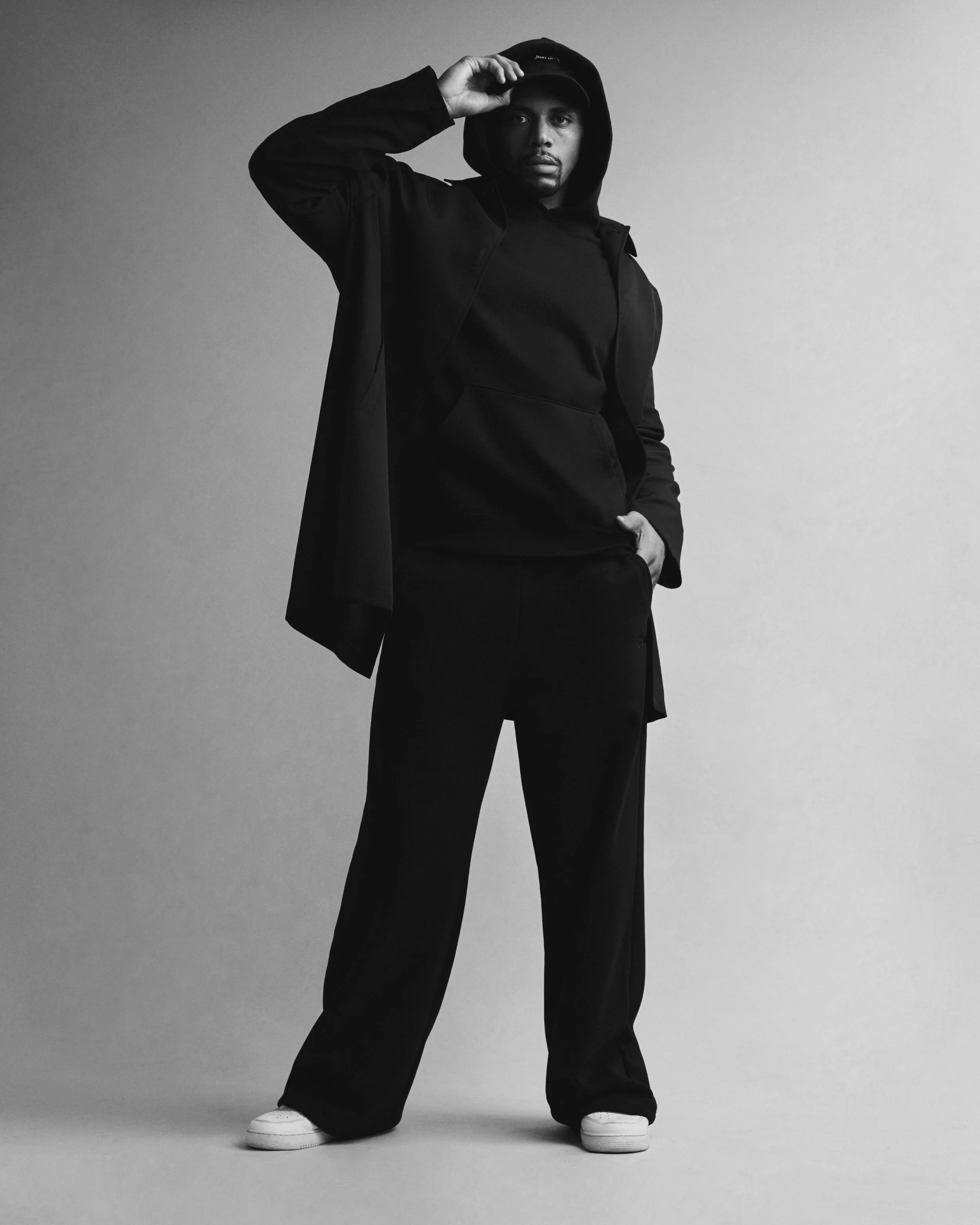A man dressed in black clothing including hoodie, pants, and a cap, standing against a plain light background. He is holding the brim of his cap with one hand and looking directly at the camera.