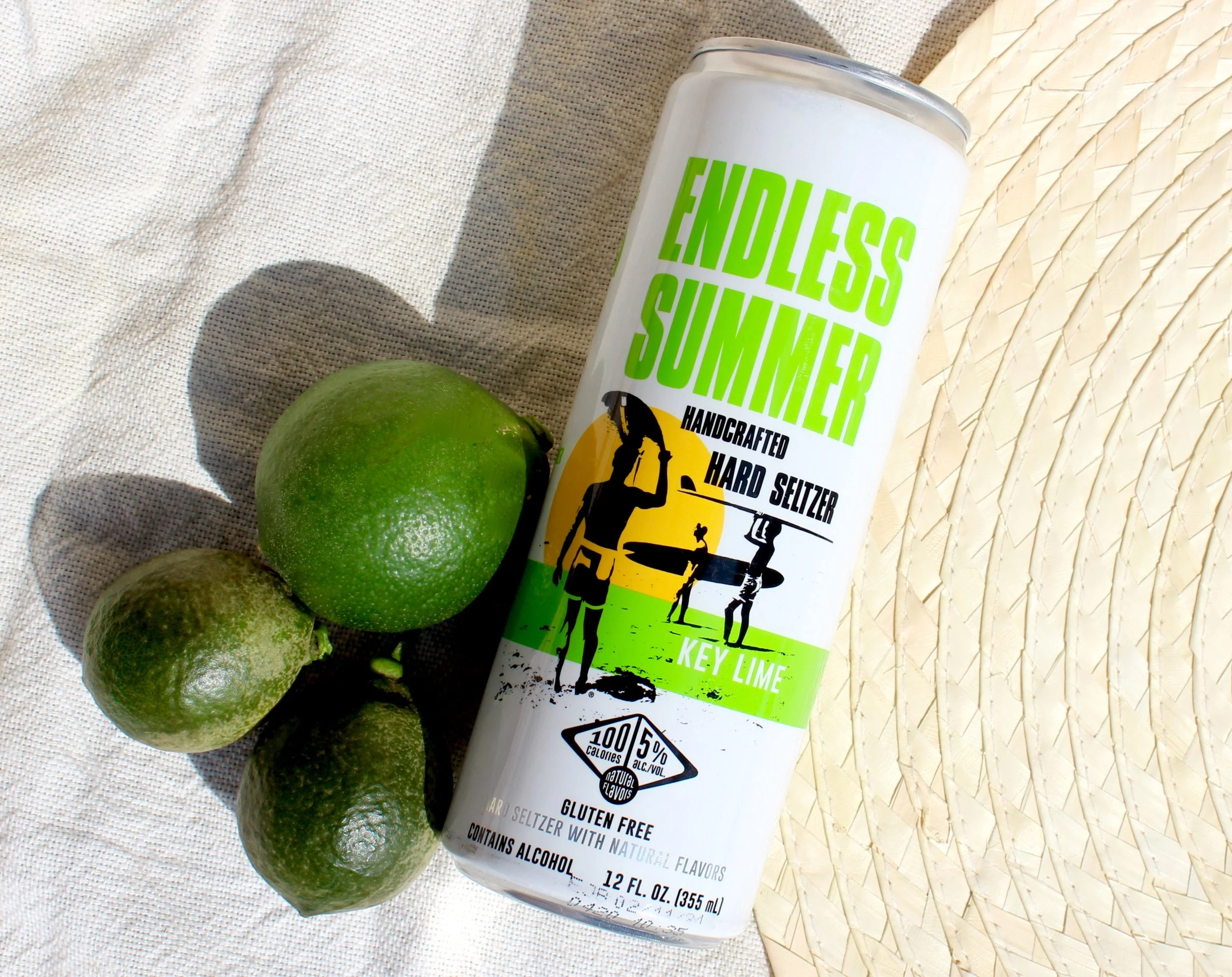 A can of Endless Summer Key Lime hard seltzer placed on a white woven surface next to three limes.