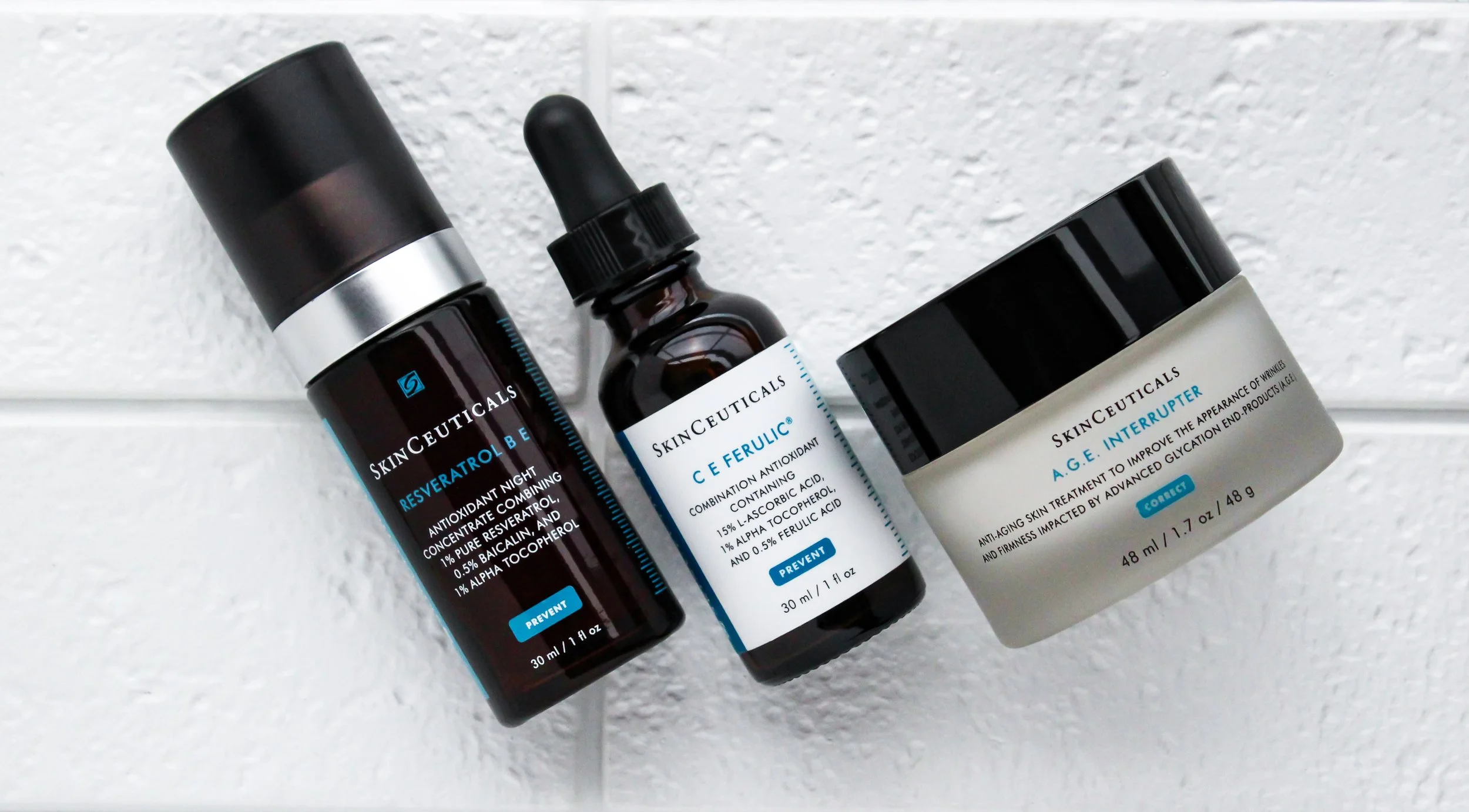 Three skincare product bottles from SkinCeuticals are placed against a white textured wall. The left bottle is black with a silver band, labeled 'Resveratrol B E,' the middle bottle is dark amber with a droplet cap labeled 'C E Ferulic,' and the righ
