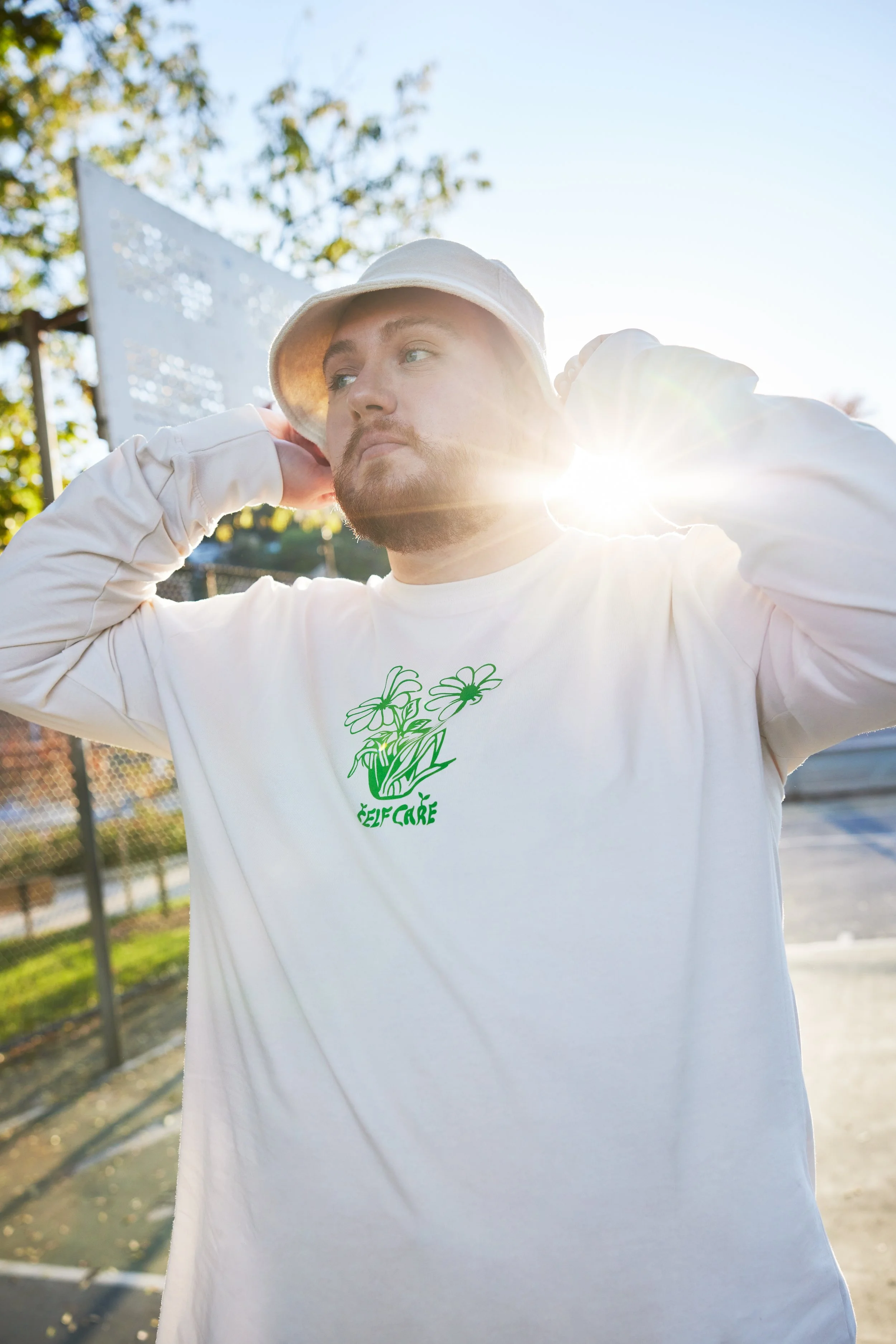 A man with a beard and blue eyes wearing a white bucket hat and white long-sleeve shirt with green floral text on the front, outdoors in a park or playground area, with trees and a chain-link fence in the background, backlit by sunlight.