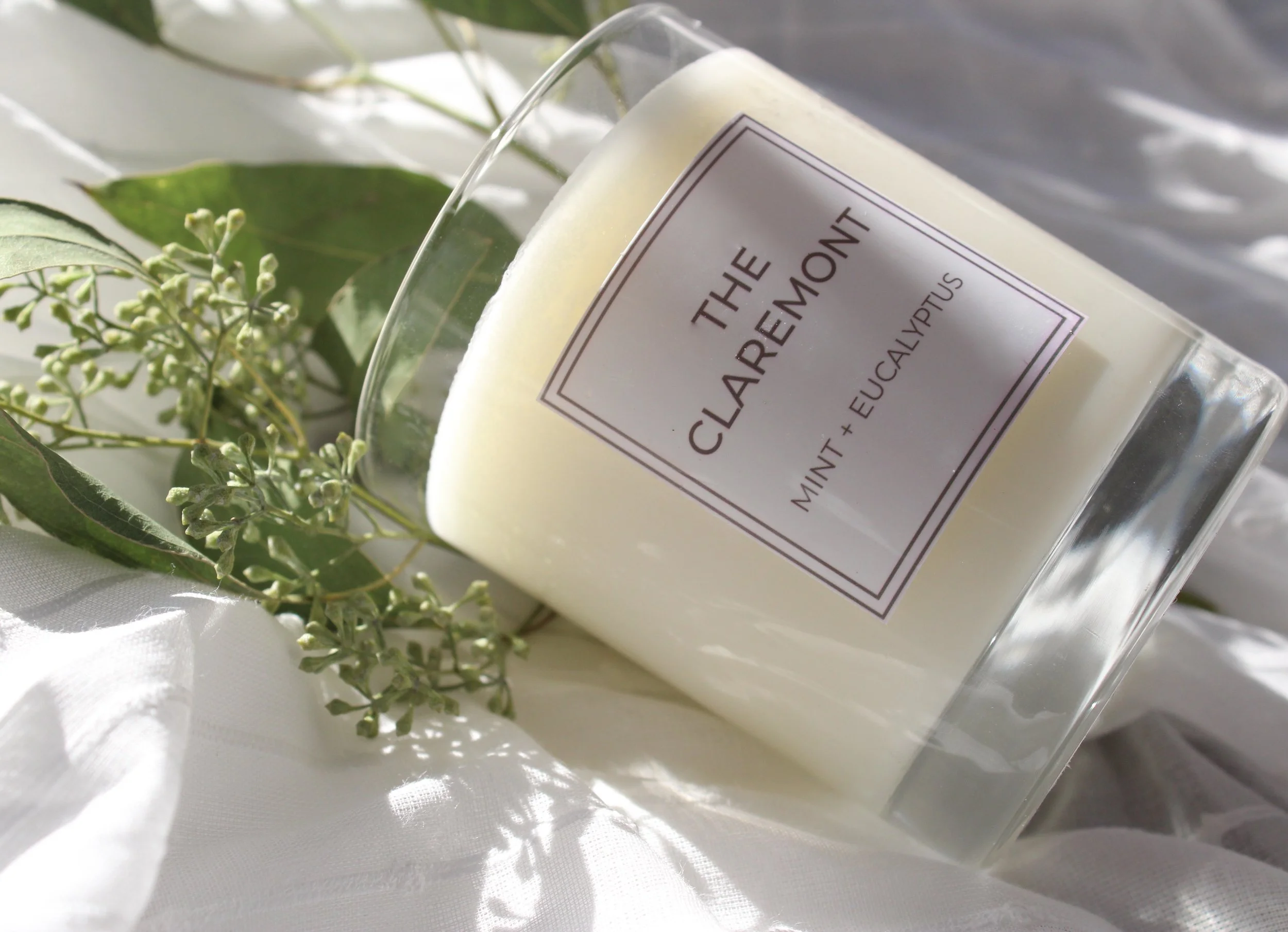 A white candle in a clear glass container with the label 'The Clarity Candle Mint + Eucalyptus', surrounded by eucalyptus leaves and small white flowers, on a white fabric surface.