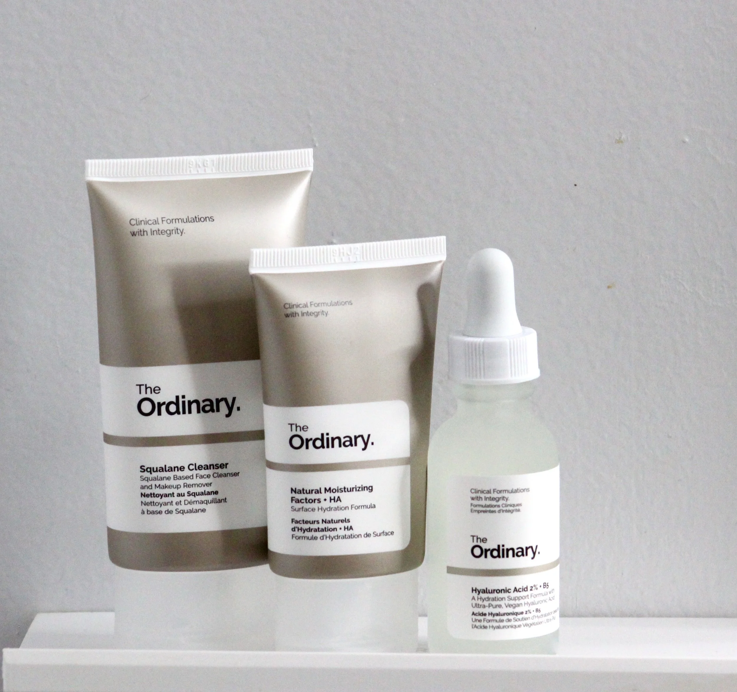 Close-up of three white tubes of skincare products with black text, placed on a white surface against a gray background. The tubes are labeled with different product names and descriptions.