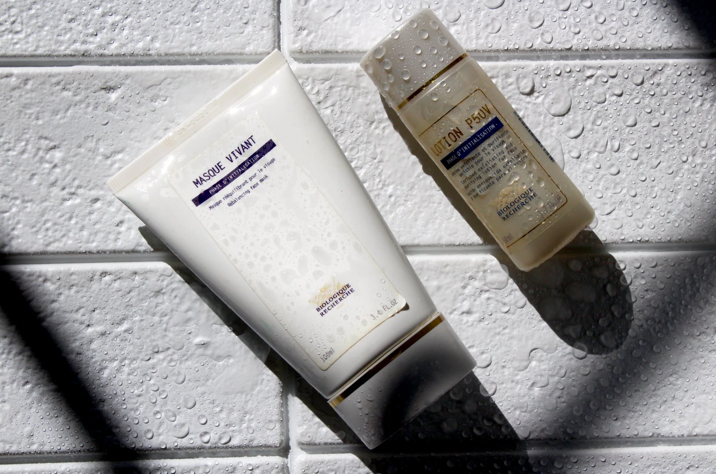 Two bottles of skincare products, one labeled 'Masque Vivant' and the other labeled 'Lotion d'Intégration,' on a wet white surface with water droplets.