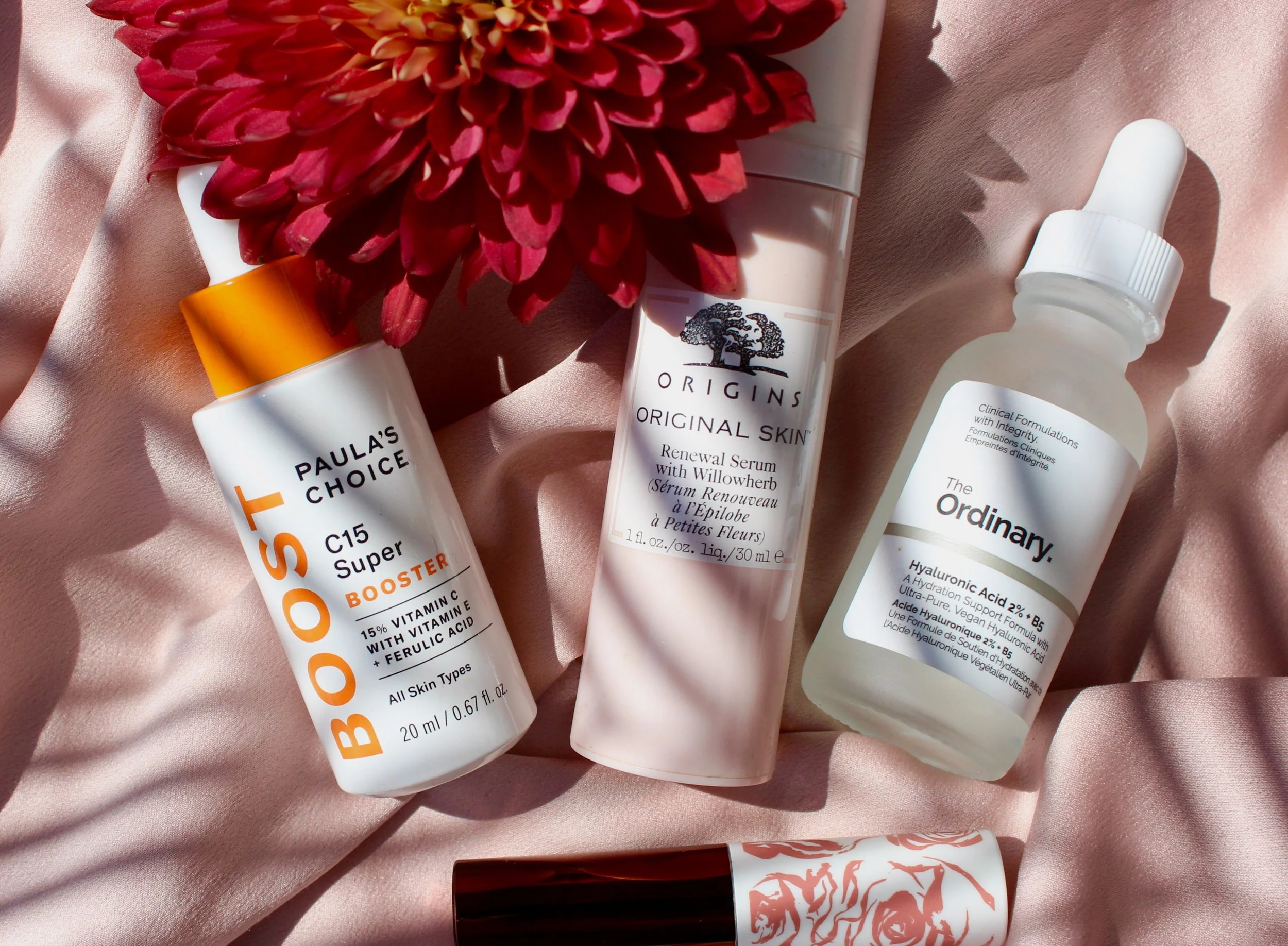 Close-up of skincare products including bottles of 'Paula's Choice,' 'Origins,' and 'The Ordinary,' arranged on a pink satin fabric with a large red flower in the background.