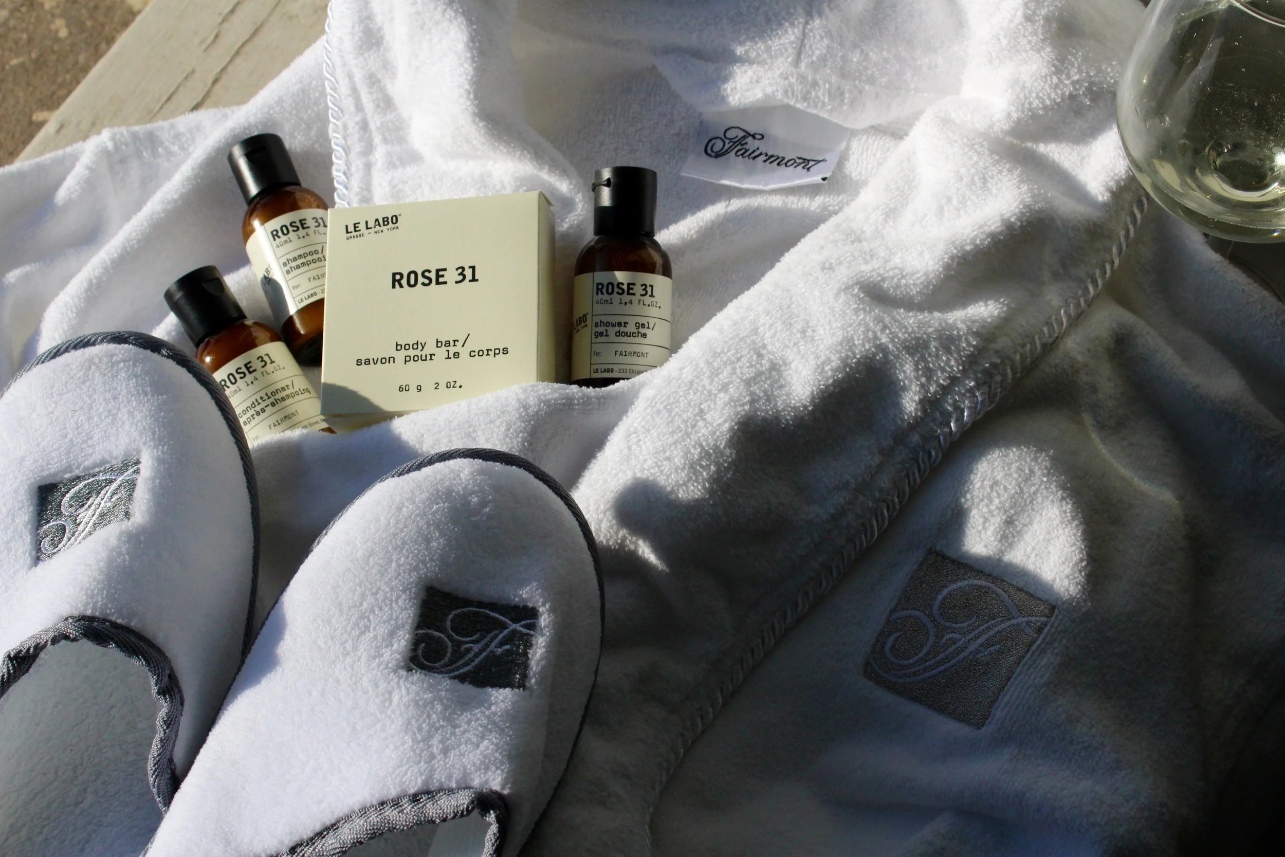 Person wearing grey sneakers with black laces, sitting on a white towel with bottles of Rose 31 body products, a bar soap, and a shower gel, along with a glass containing a clear beverage, on a wooden surface outdoors.