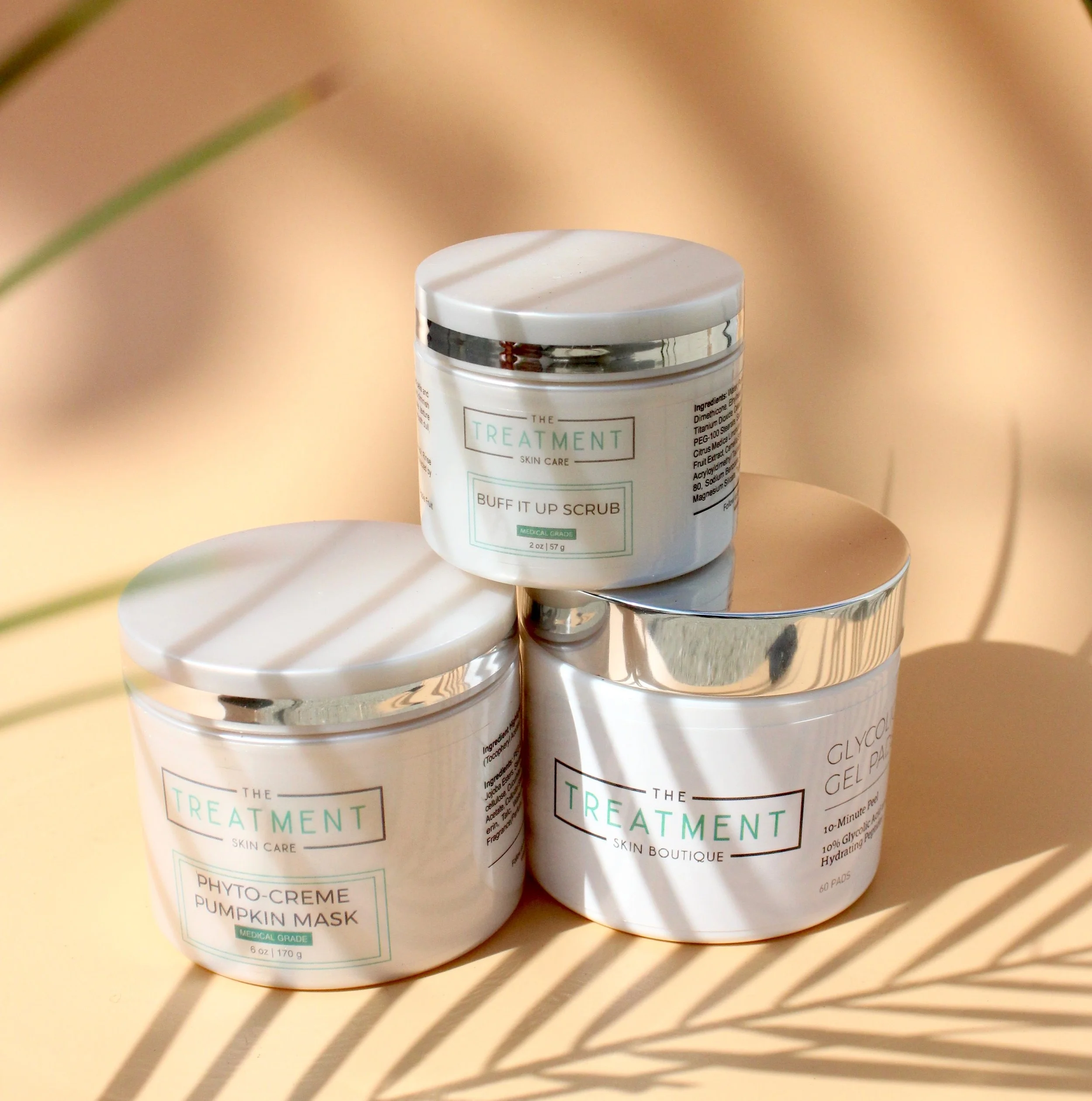 Three white jars of skincare products on a reflective metallic surface, partially obscured by shadows from a patterned decorative object, with a blurred beige background.