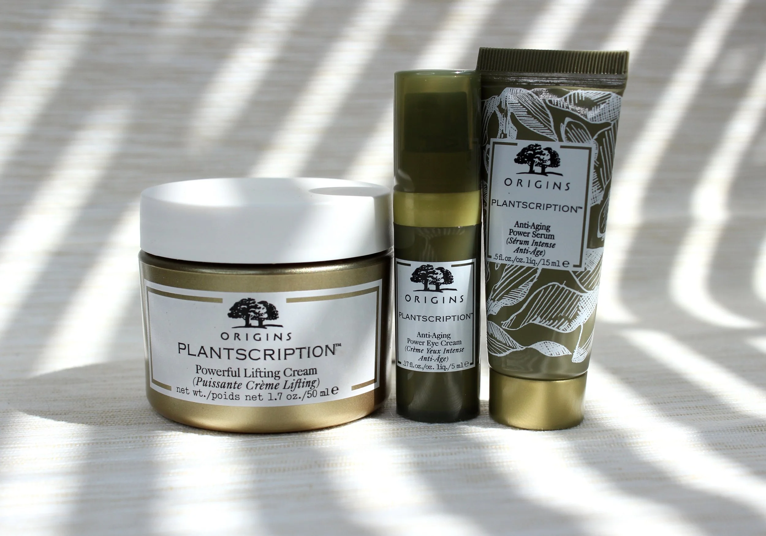 Three skincare products from the Origins Plantscription line, including a powerful lifting cream, an anti-aging power serum, and an anti-aging power eye cream, placed on a surface with bright sunlight and shadows.
