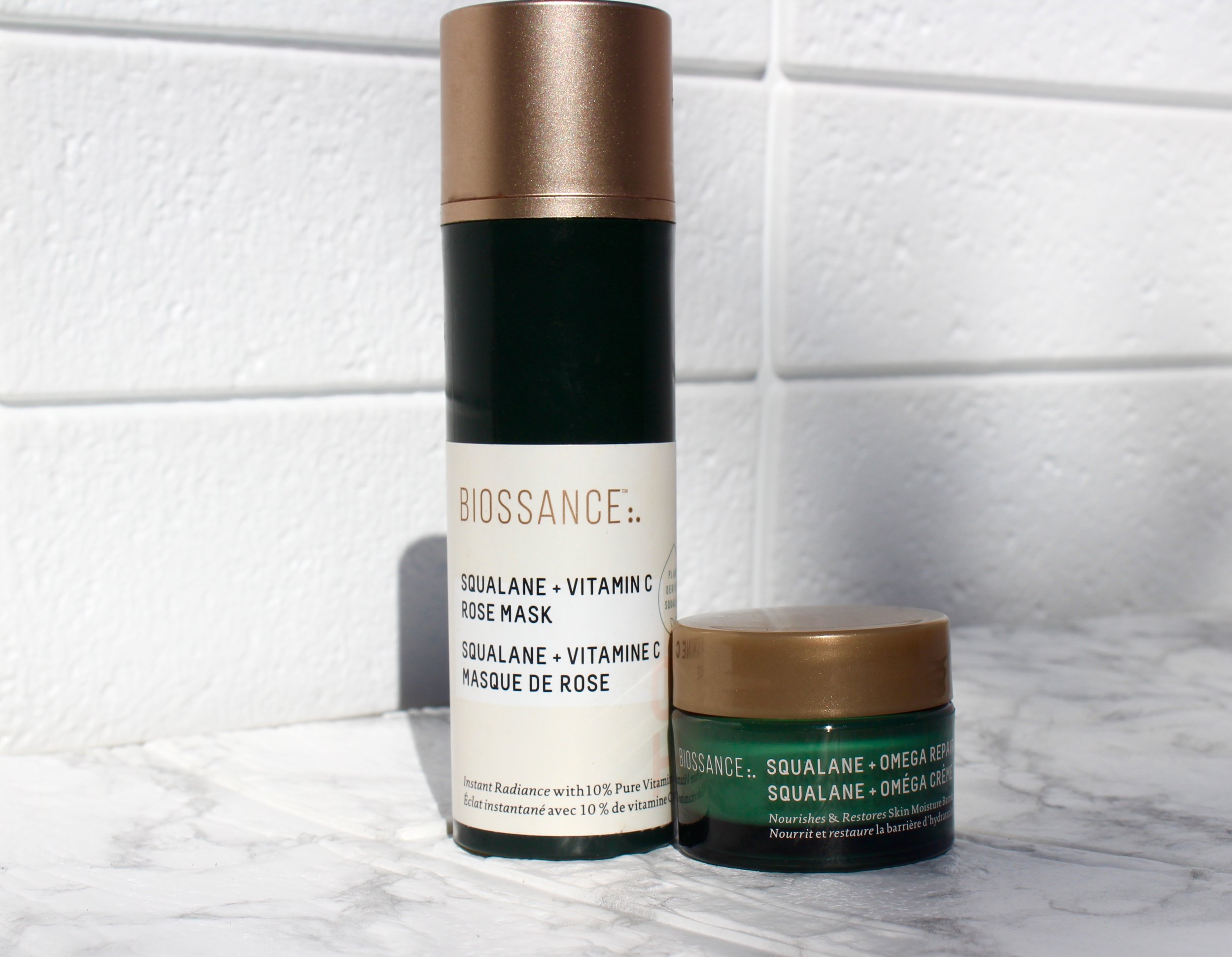 Three skincare product bottles from the Biosance brand, positioned against a white tiled background. The bottles feature white, black, and teal colors with gold accents and contain labels with product descriptions.