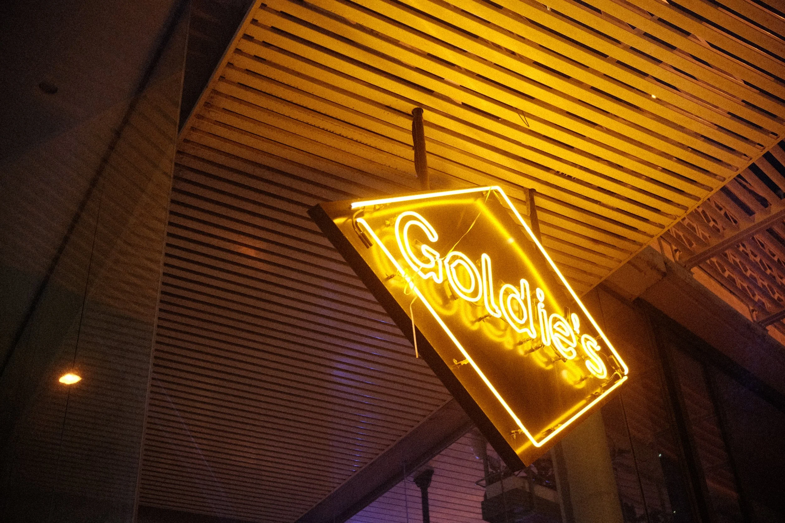 A neon sign with the word 'Goldies' in yellow, hanging under a ceiling with wooden slats at night.