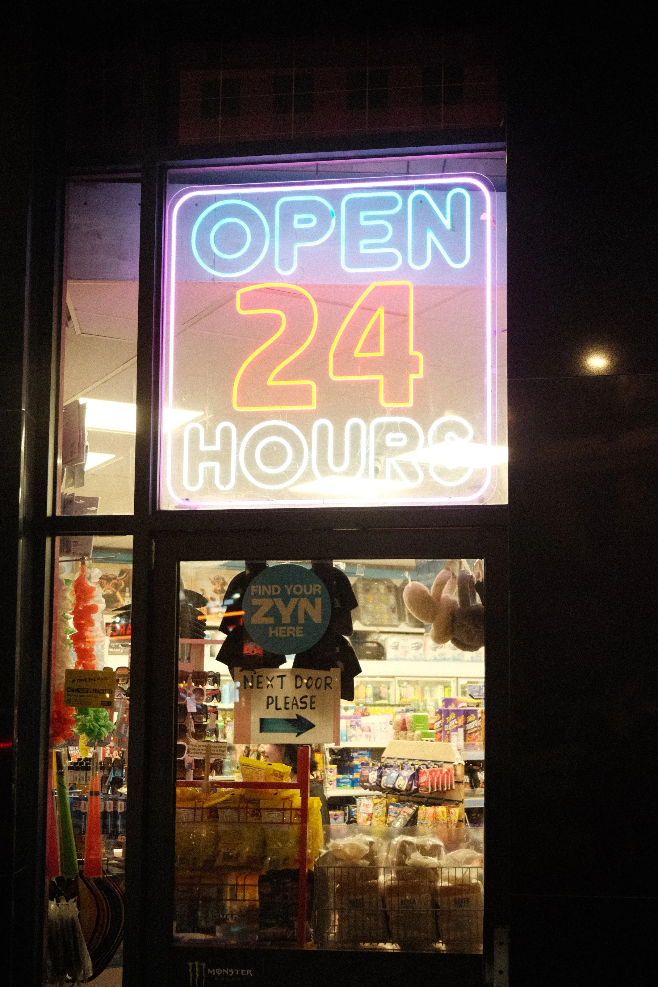 Night view of a convenience or retail store with neon sign that reads 'Open 24 Hours' in bright neon colors. The store window displays various products and signs inside, including a sign that says 'Find Your ZYN Here' and a 'Next Door Please' sign wi