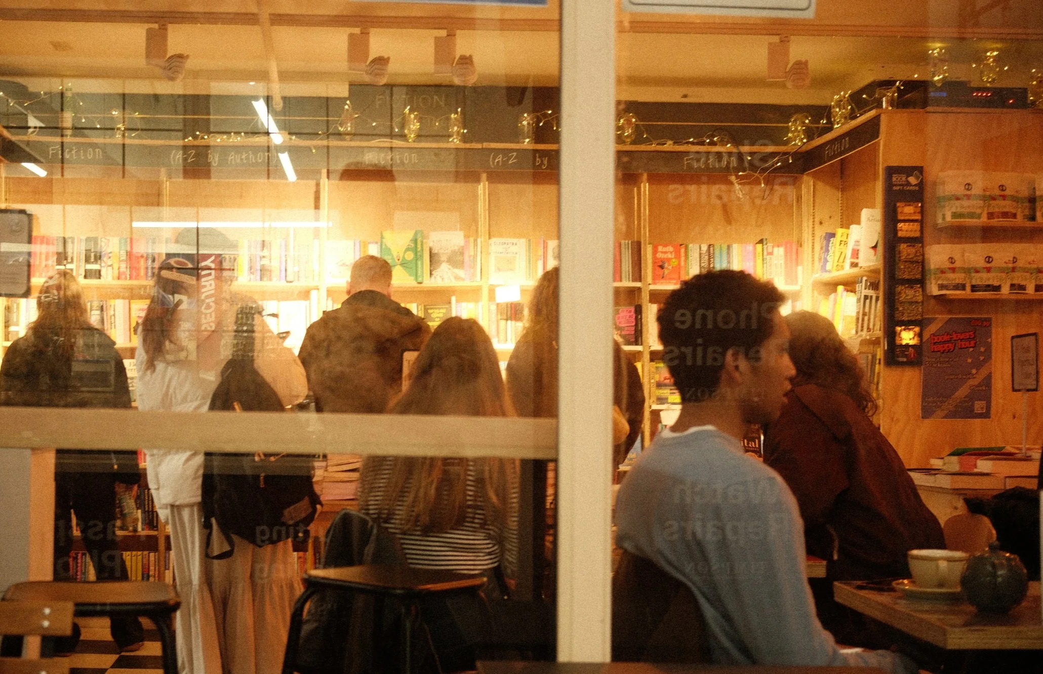 Inside a bookstore or cafe with wooden shelves filled with books and warm lighting, where several people are standing and sitting, engaged in conversation or browsing.