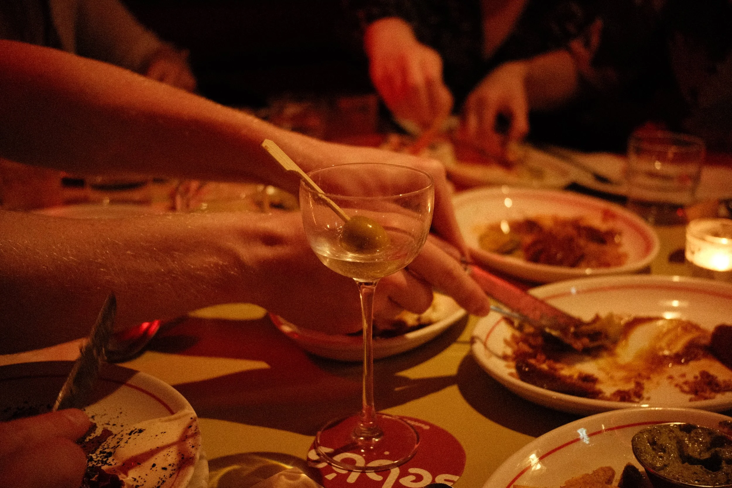 A person reaching for food at a dimly lit table with various plates of appetizers and a drink in the foreground.