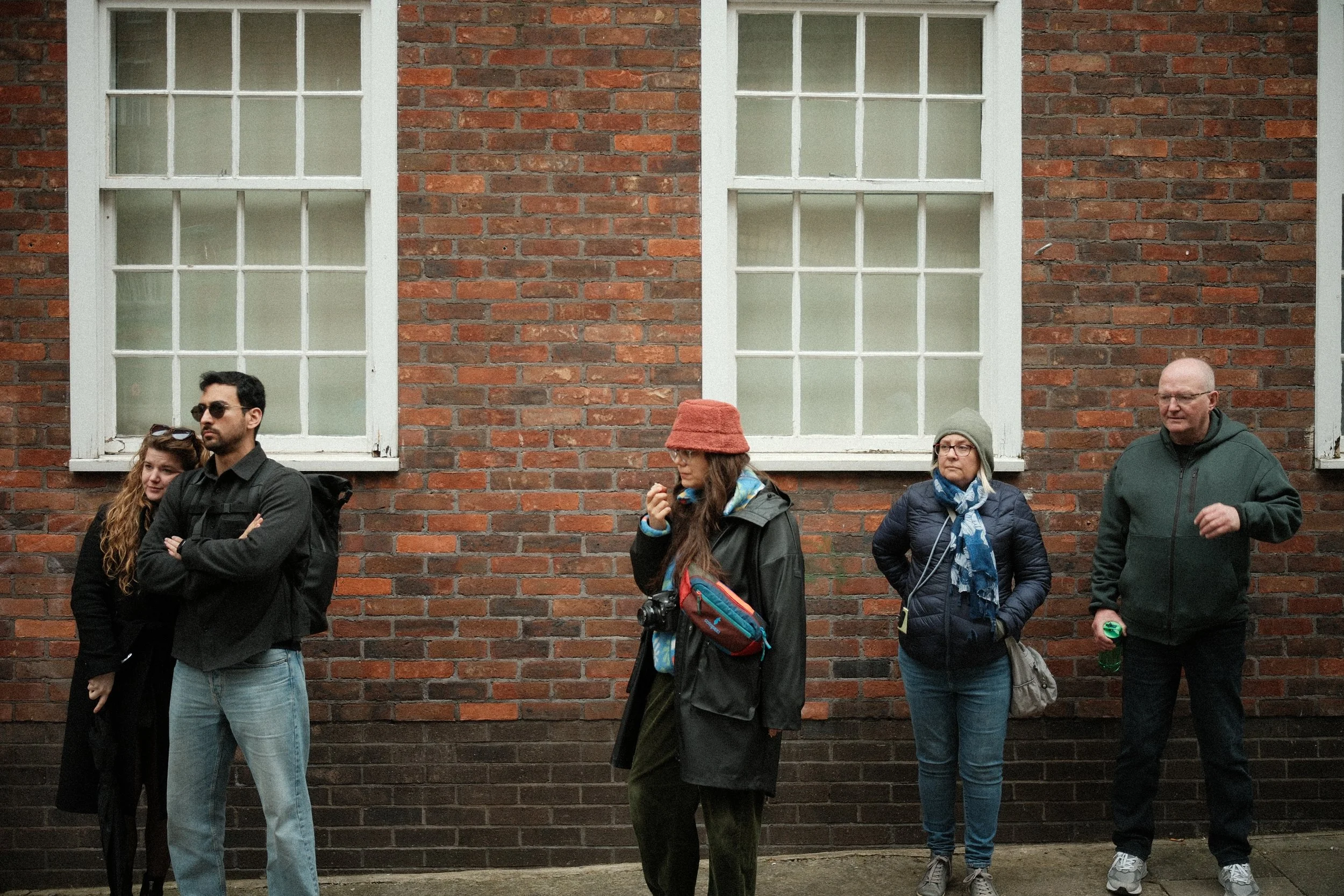 Group of five people standing against a brick wall with two large windows, some wearing jackets and carrying backpacks, one with sunglasses, and one holding a water bottle.