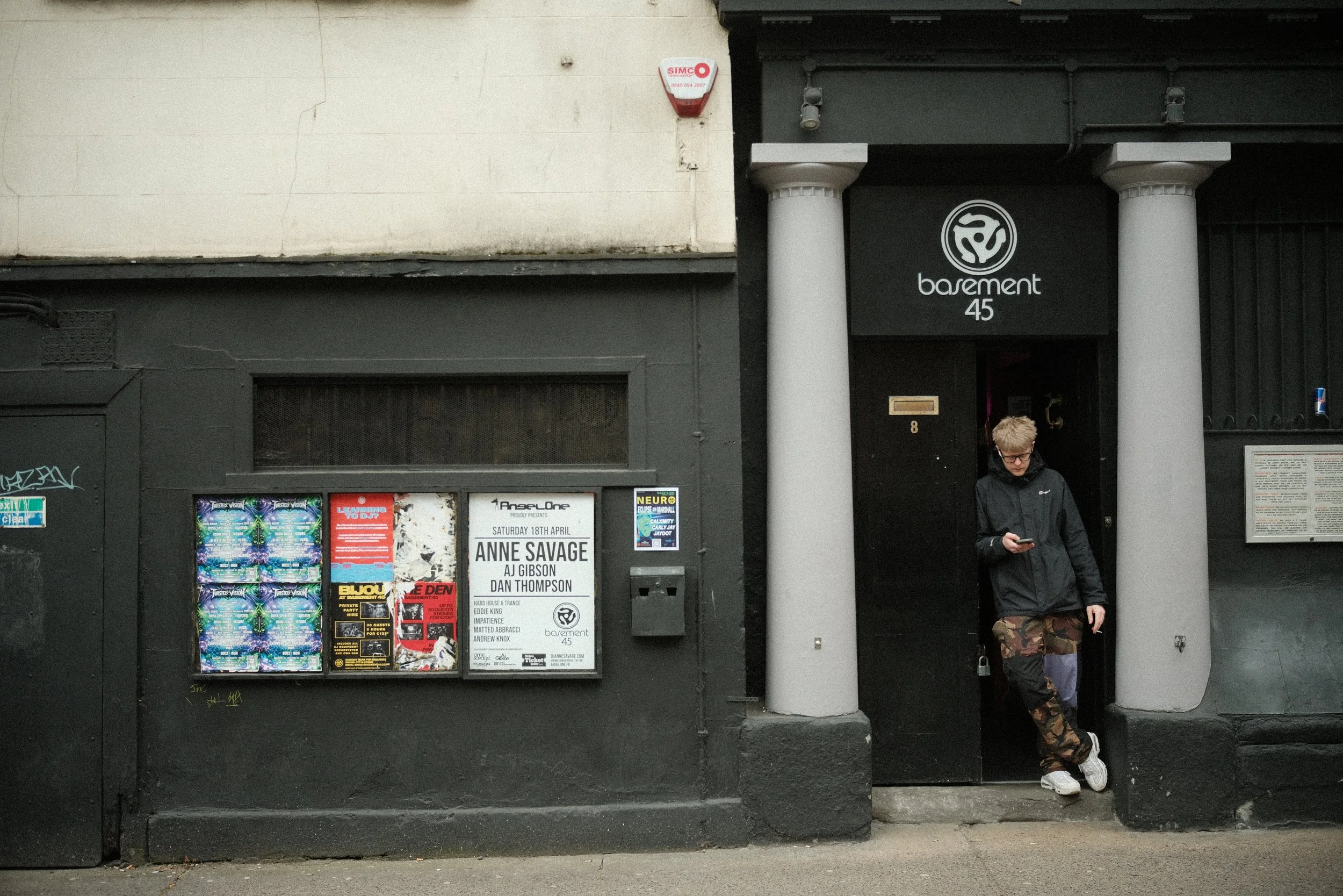 A young man with blond hair and glasses, wearing a black jacket and camouflage pants, standing on the sidewalk next to a black building with white pillars and a sign that reads 'basement 45'. There are posters on the window, and a small window with g