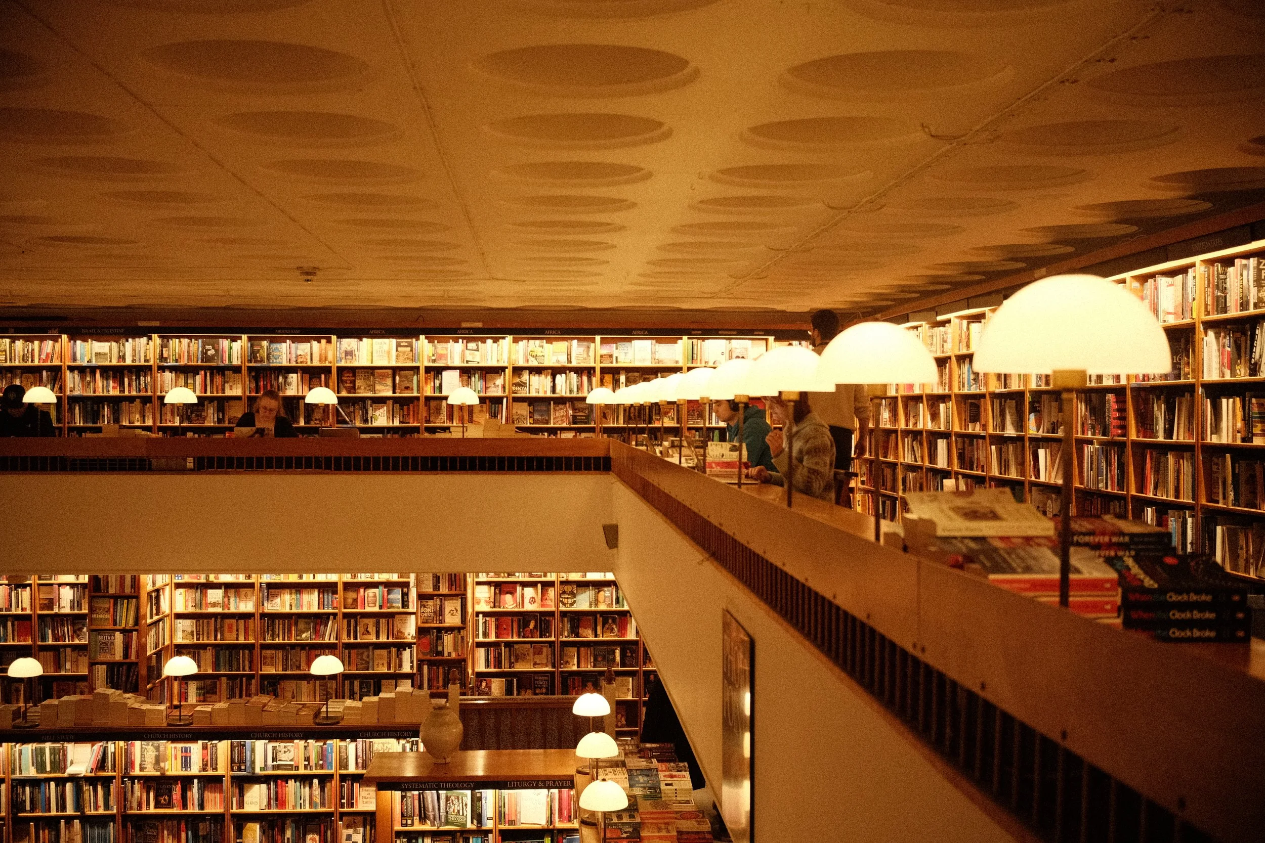 Bookshelves filled with books in a library, illuminated by hanging lamps, with a person reading at a table.