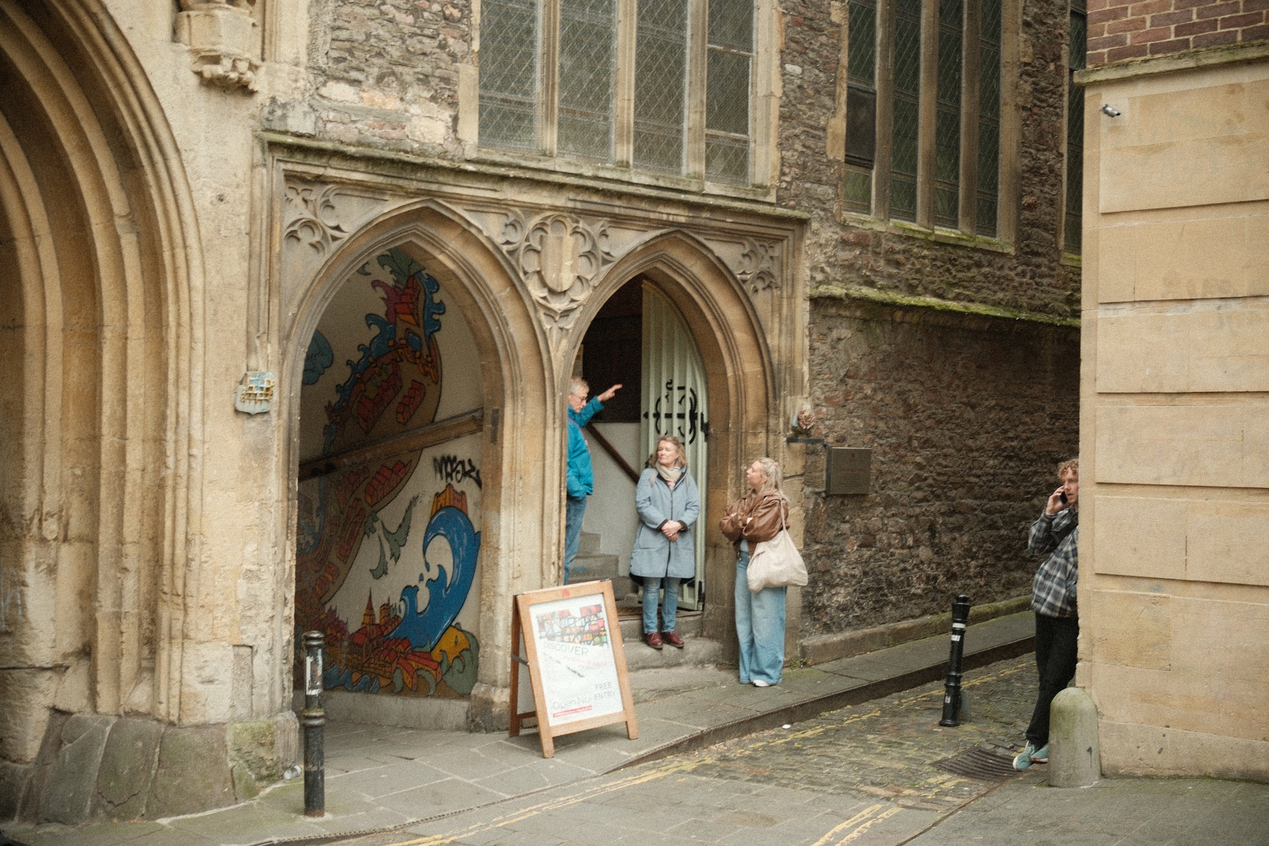 People standing outside a historic stone building with arched entrance and stained glass windows, some talking and one taking a photo.