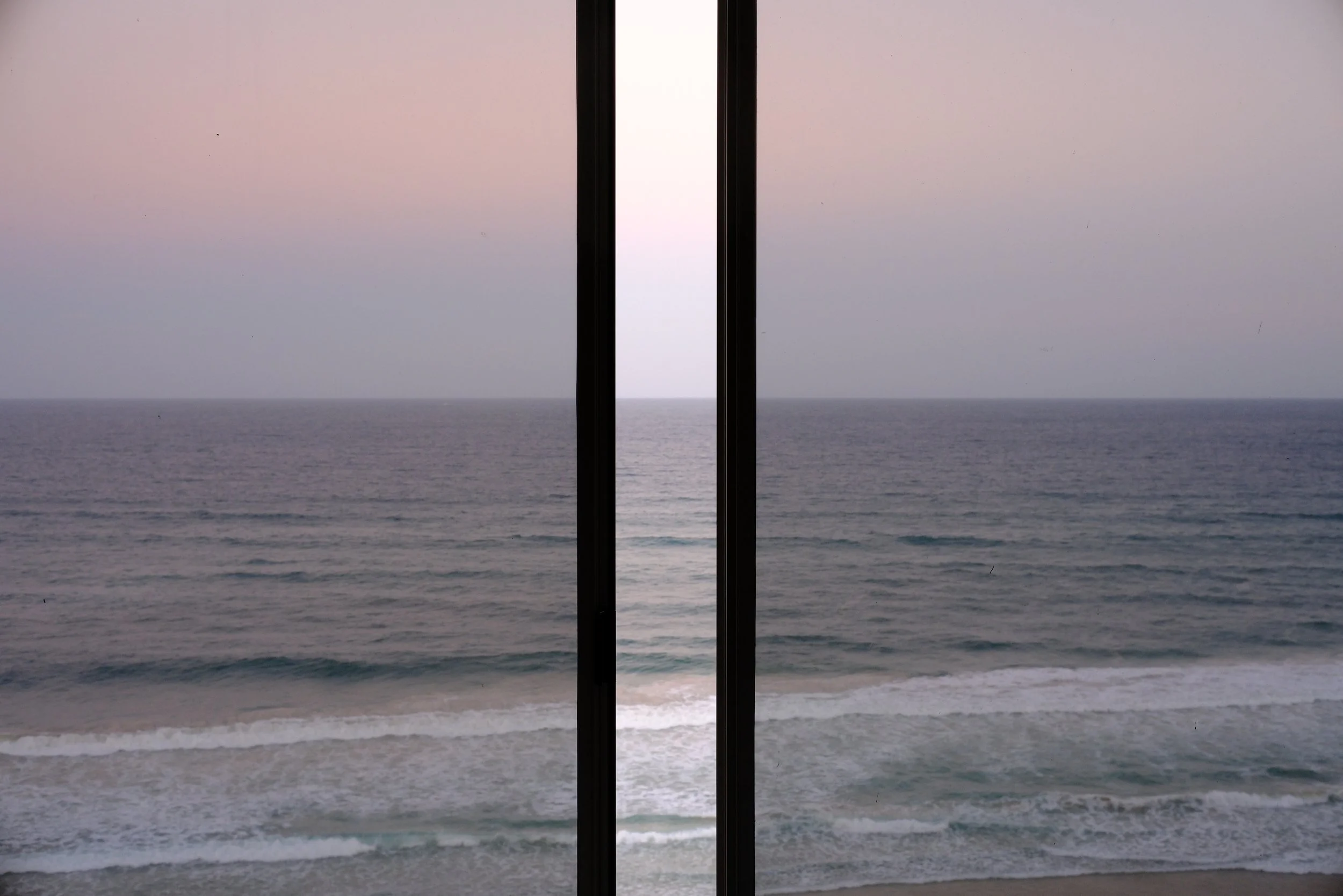 View of the ocean seen through glass doors, with waves gently crashing on the beach, under a pale sky at sunset or sunrise.