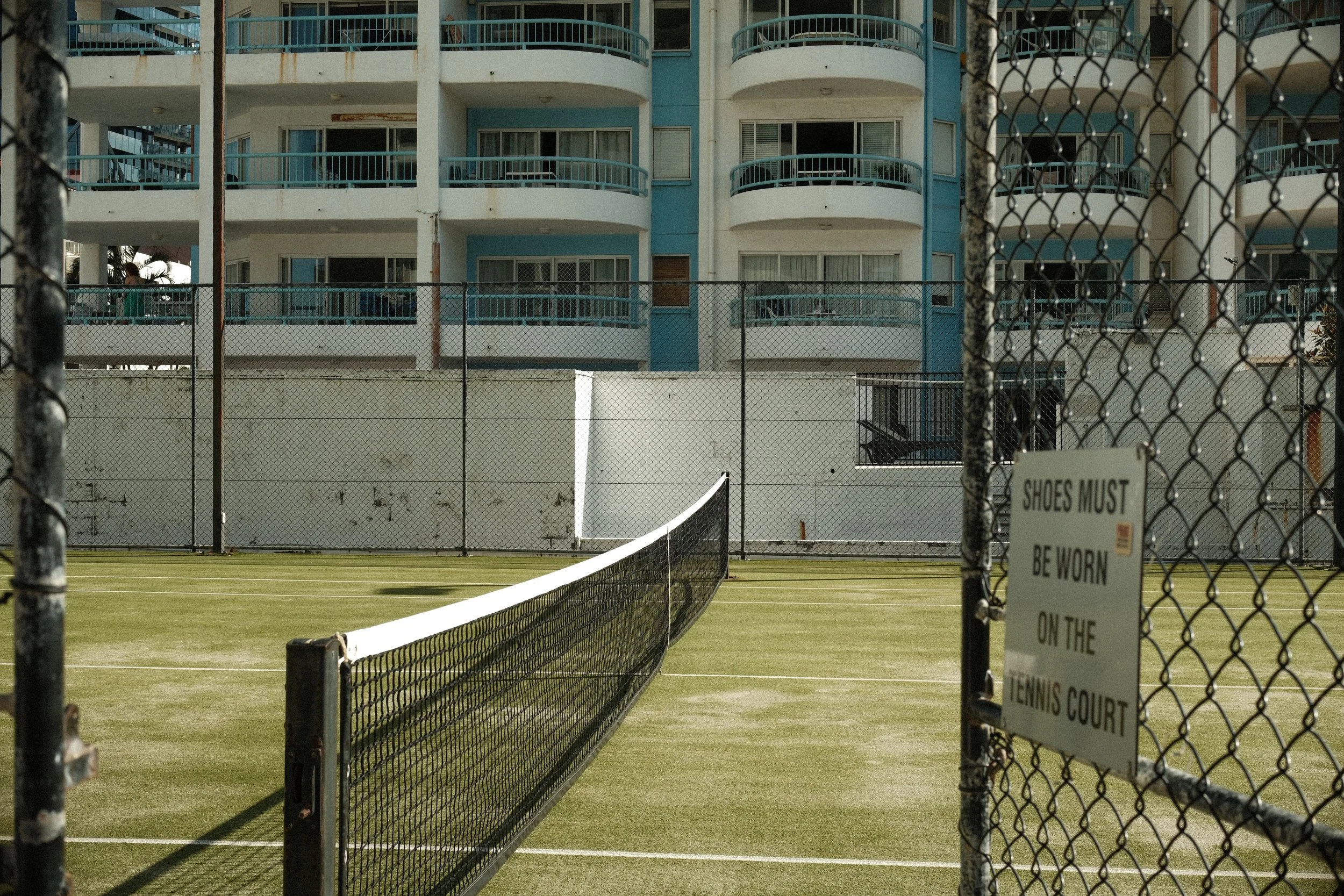 A fenced tennis court with a sign that reads 'SHOES MUST BE WORN ON THE TENNIS COURT', surrounded by a multi-story residential or hotel building with balconies and windows.