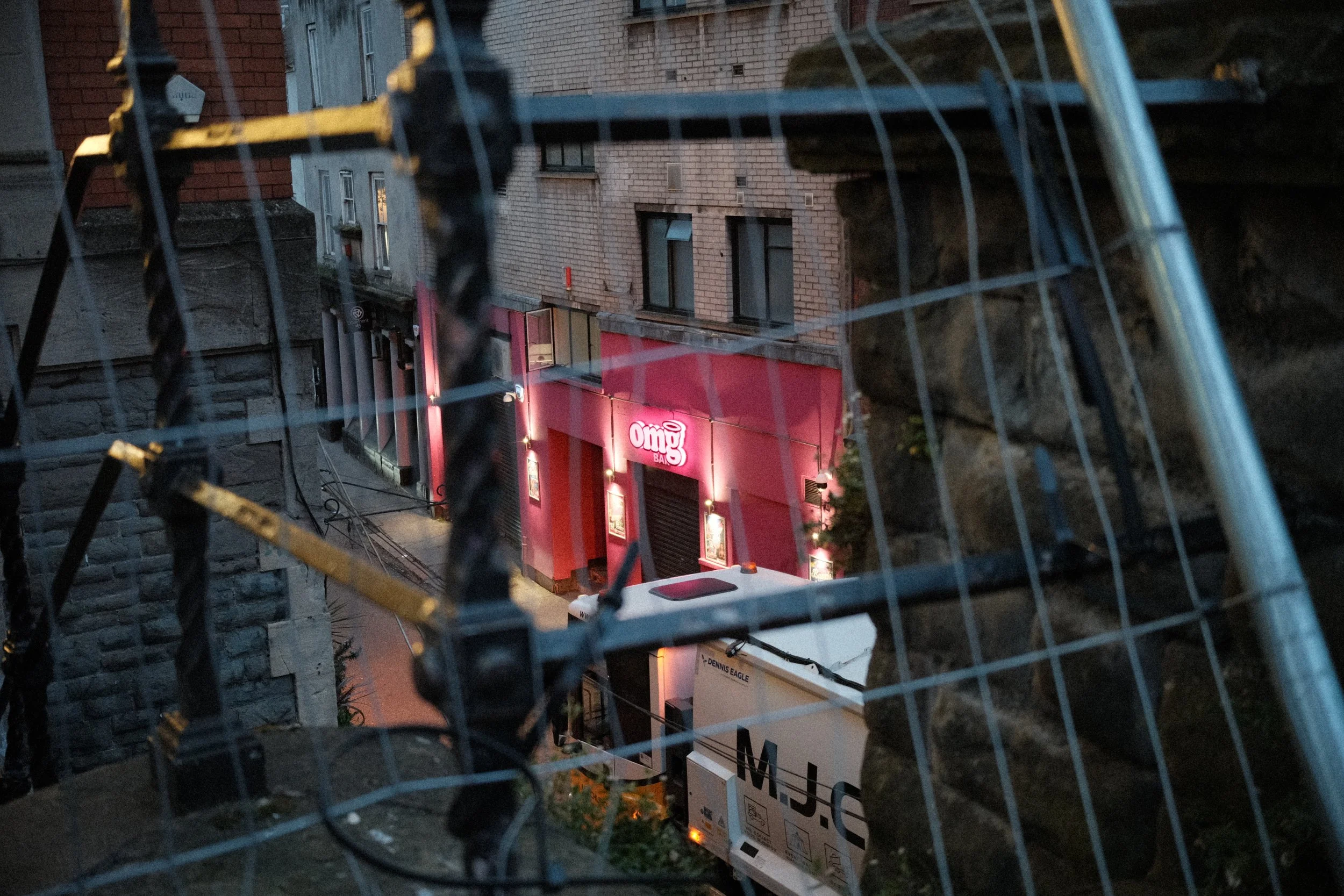 Urban alleyway viewed through a chain-link fence at dusk. Buildings with brick and stone facades line the alley, and a pink storefront sign reads 'Omg'.