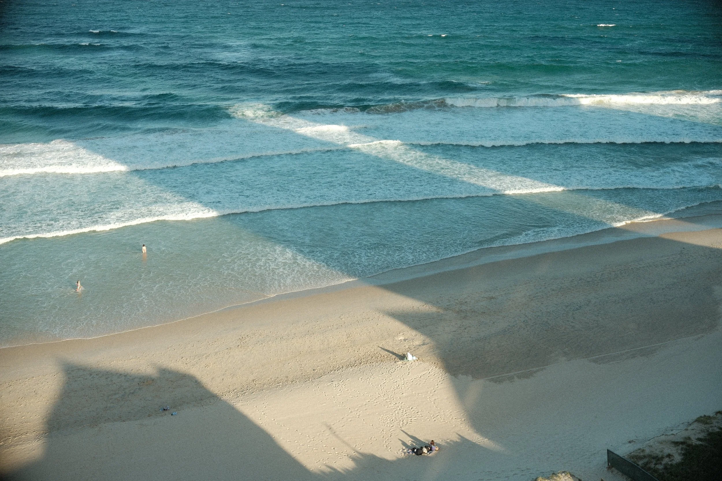 View of a sandy beach with people, ocean waves, and a shadow from a bridge or structure overhead.