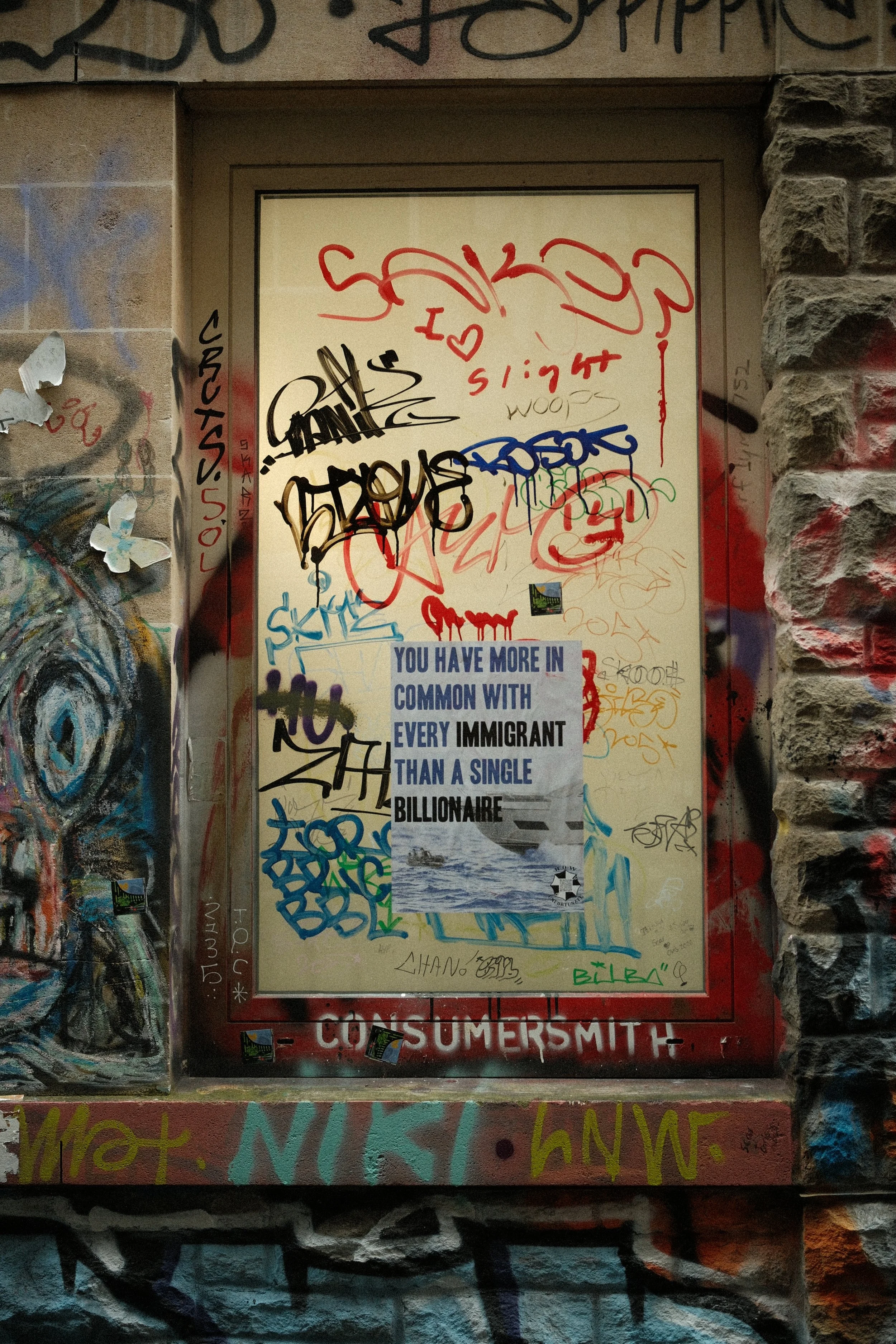 Graffiti-covered wall with stickers, and a poster that reads, 'You have more in common with every immigrant than a single billionaire.'