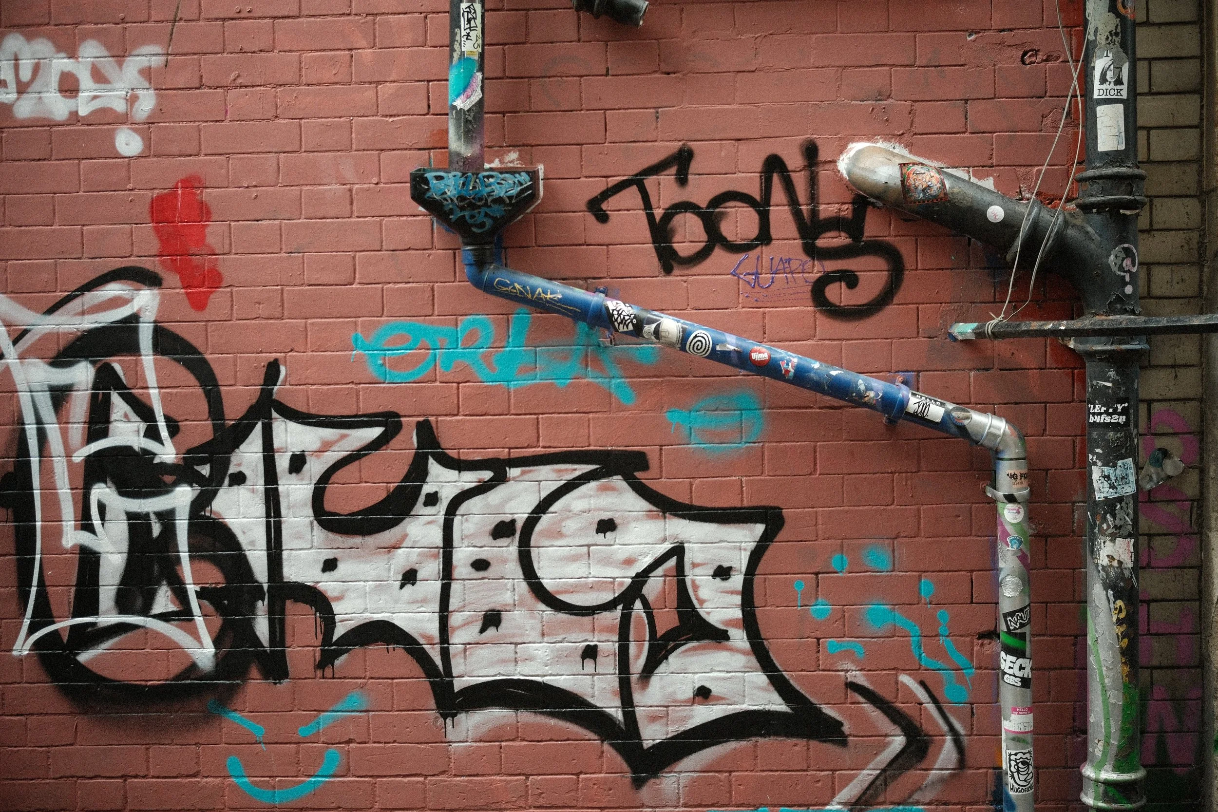 A brick wall covered in various graffiti tags, with a metal pipe running horizontally across the wall, adorned with stickers and graffiti.