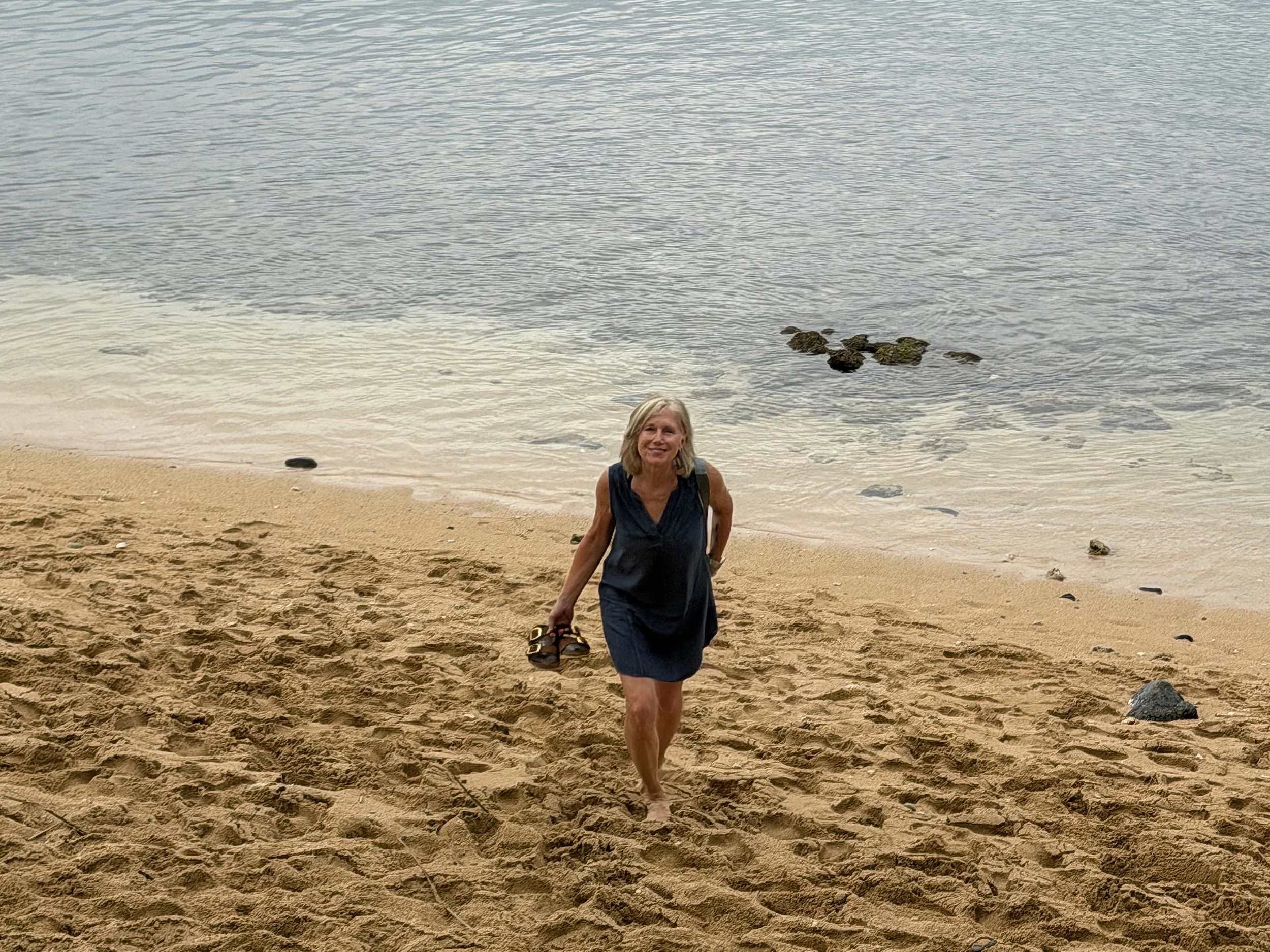 Seattle life coach Paula Freeman of Affirmative Coaching is wearing a dark sleeveless top and a skirt, walking on a sandy beach near the water's edge, holding her shoes in her right hand, with calm blue water and some rocks in the background.