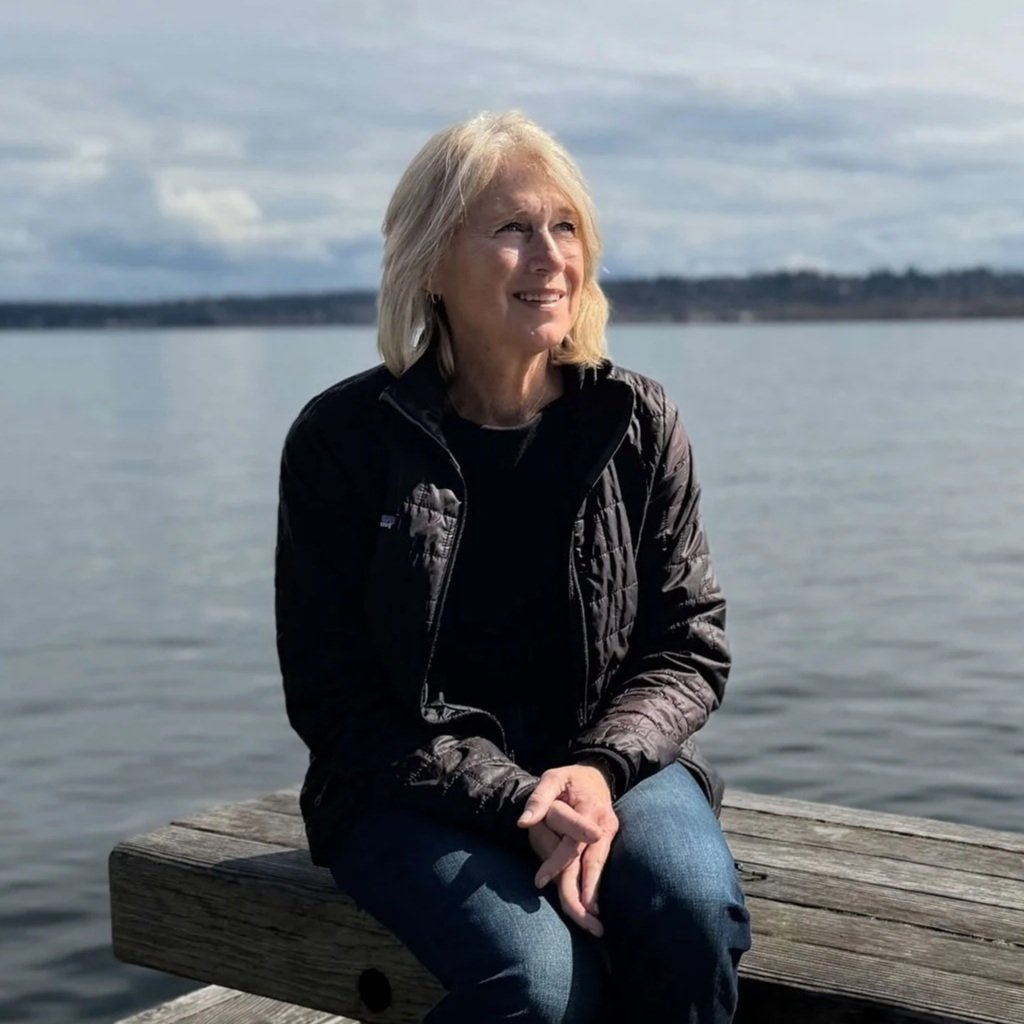 Seattle life coach Paula Freeman of Affirmative Coaching sits by a body of water, looking into the distance with a thoughtful expression, wearing a black quilted jacket and blue jeans.
