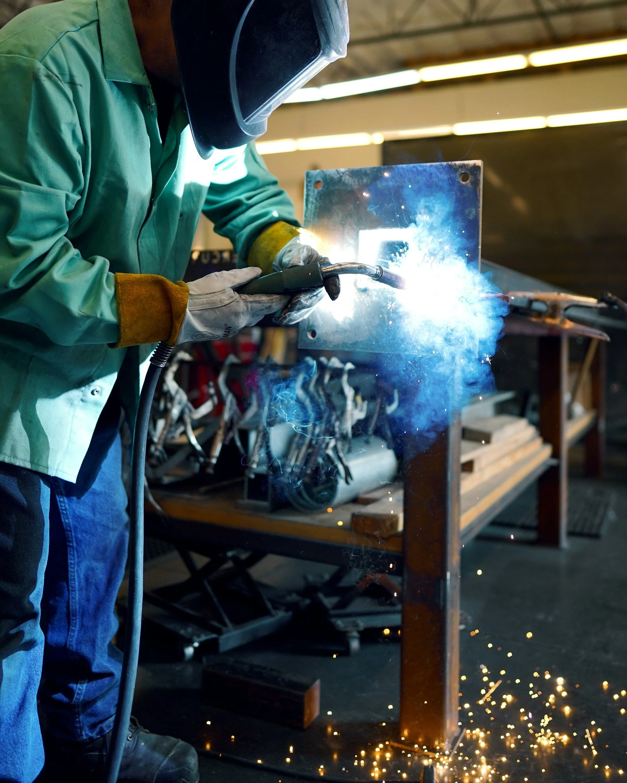 A person welding metal in a workshop, wearing protective gear including a welding helmet, green shirt, gloves, and jeans, with sparks emanating from the welding process.