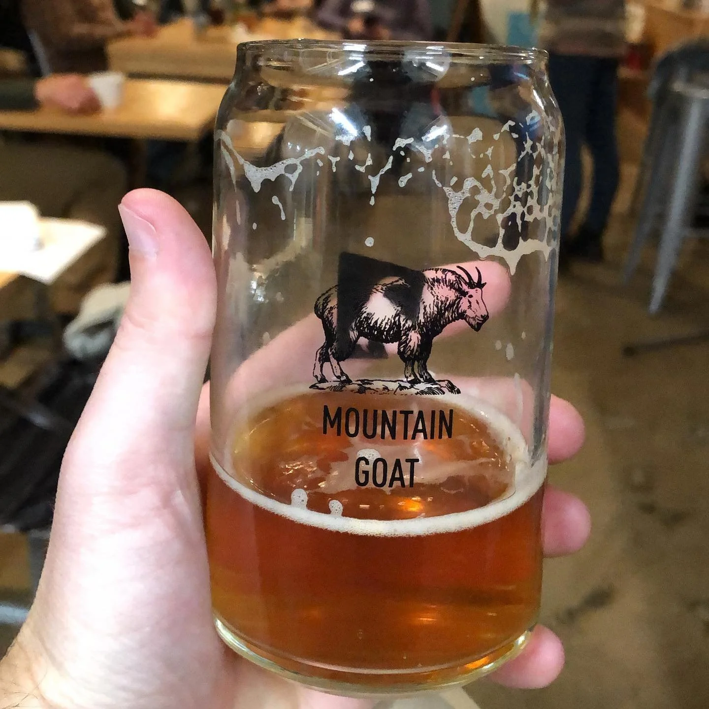 A clear glass with a mountain goat logo and the words "Mountain Goat" printed on it, containing a small amount of amber-colored beer.