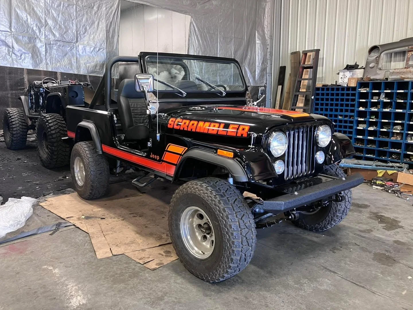 A black Jeep Scrambler with red and orange accents parked inside a workshop or garage, with one front wheel on a piece of cardboard and the other on the concrete floor. The vehicle is partially disassembled, with the rear section unfinished and various tools and parts on shelves in the background.