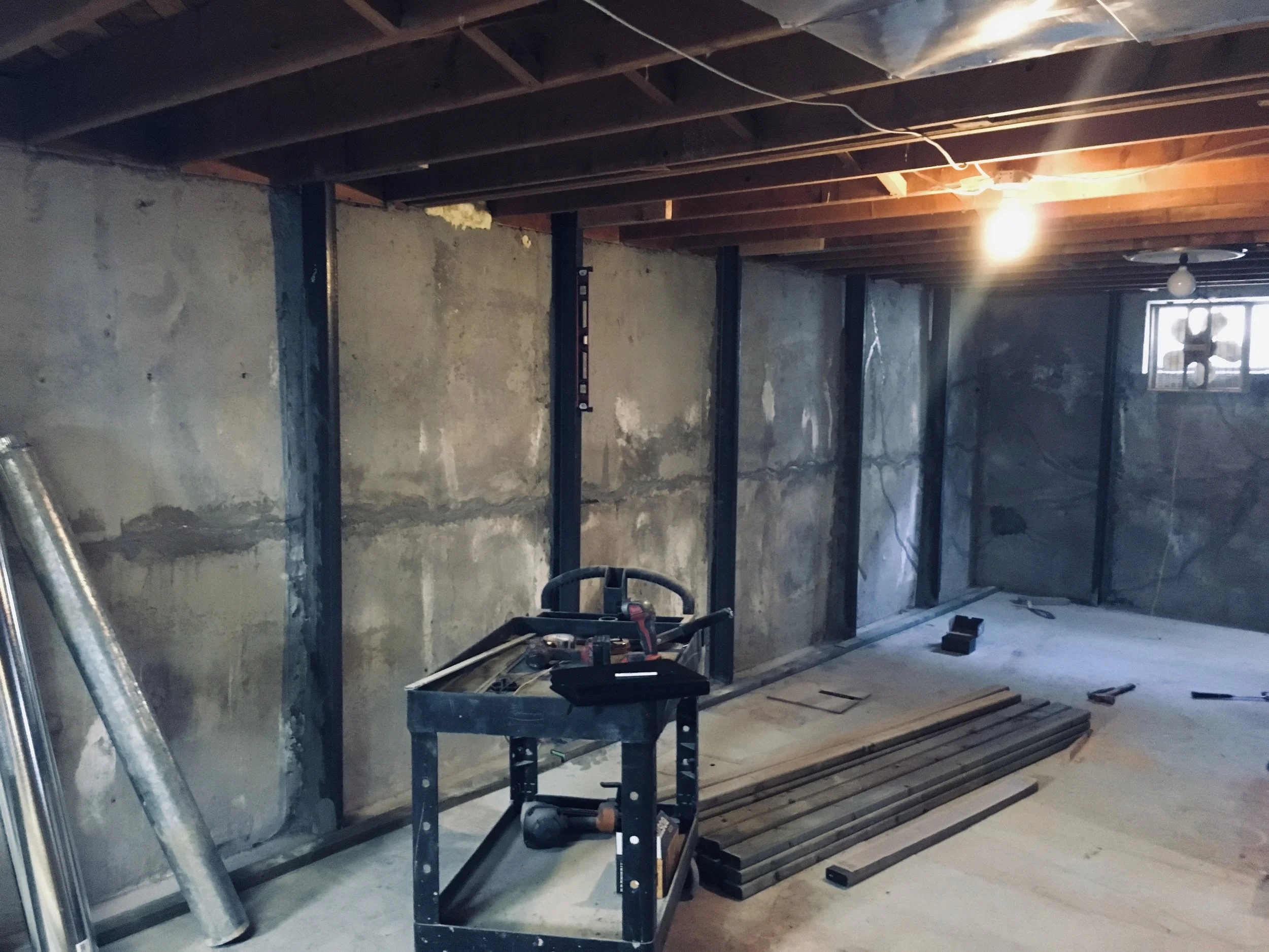 Interior view of a room under construction with exposed wooden ceiling beams, unfinished walls, construction tools and materials like metal studs and wood planks scattered on the floor, and a window at the far end.