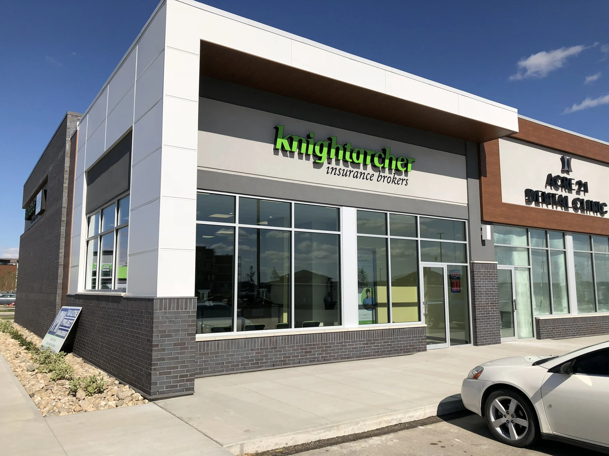 Modern commercial building with a sign for KnightArcher insurance brokers and a dental clinic. The building features large windows, white and dark brick exterior, and a sidewalk in front with parked cars.