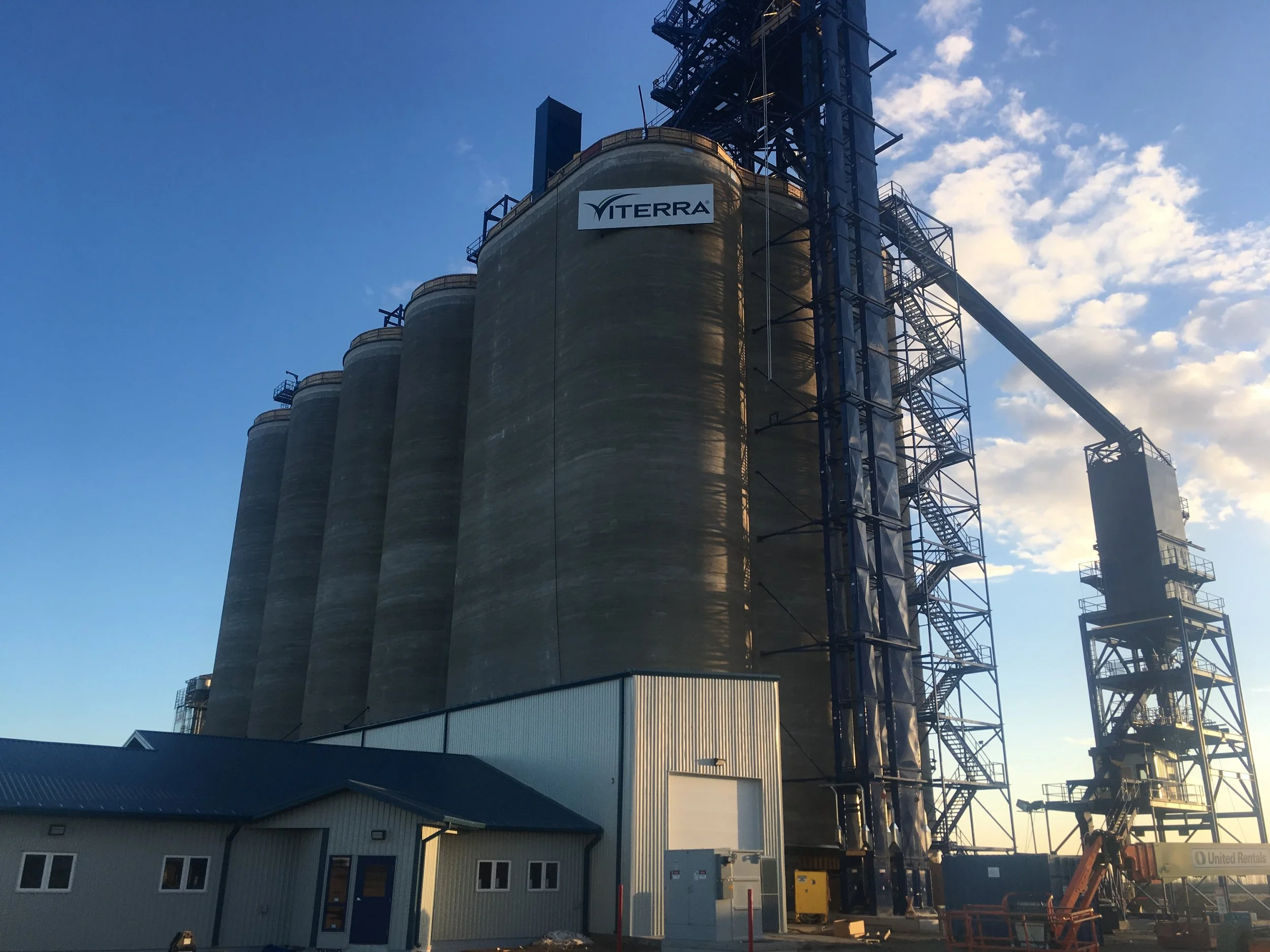 Industrial scene with a large concrete silo and a steel structure with stairs and a platform. A sign reading 'Viterra' is attached to the silo, and the sky is partly cloudy with the sun shining.