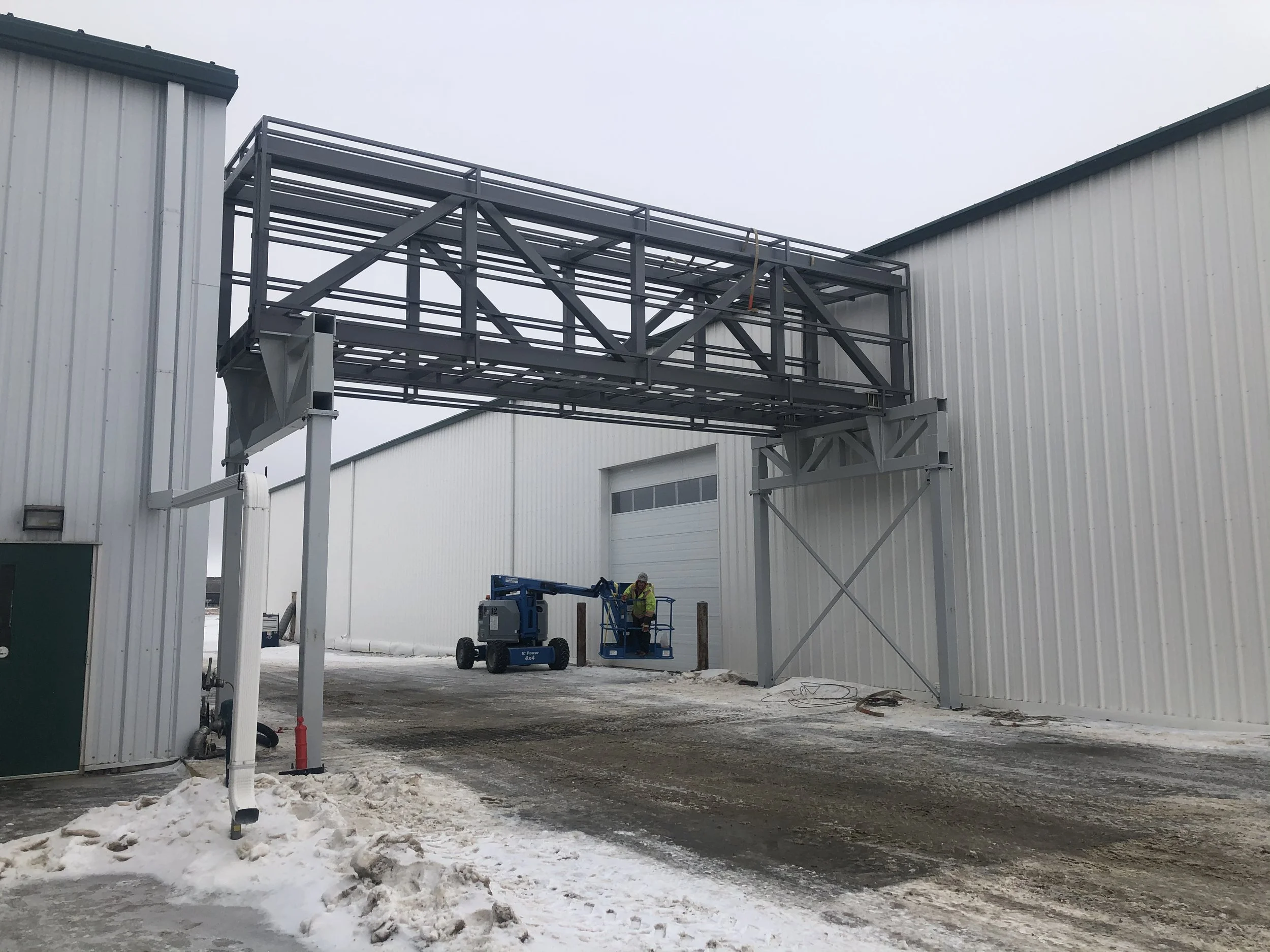 Construction workers using a lift to work on a metal framework outside a large industrial building on a snowy day.