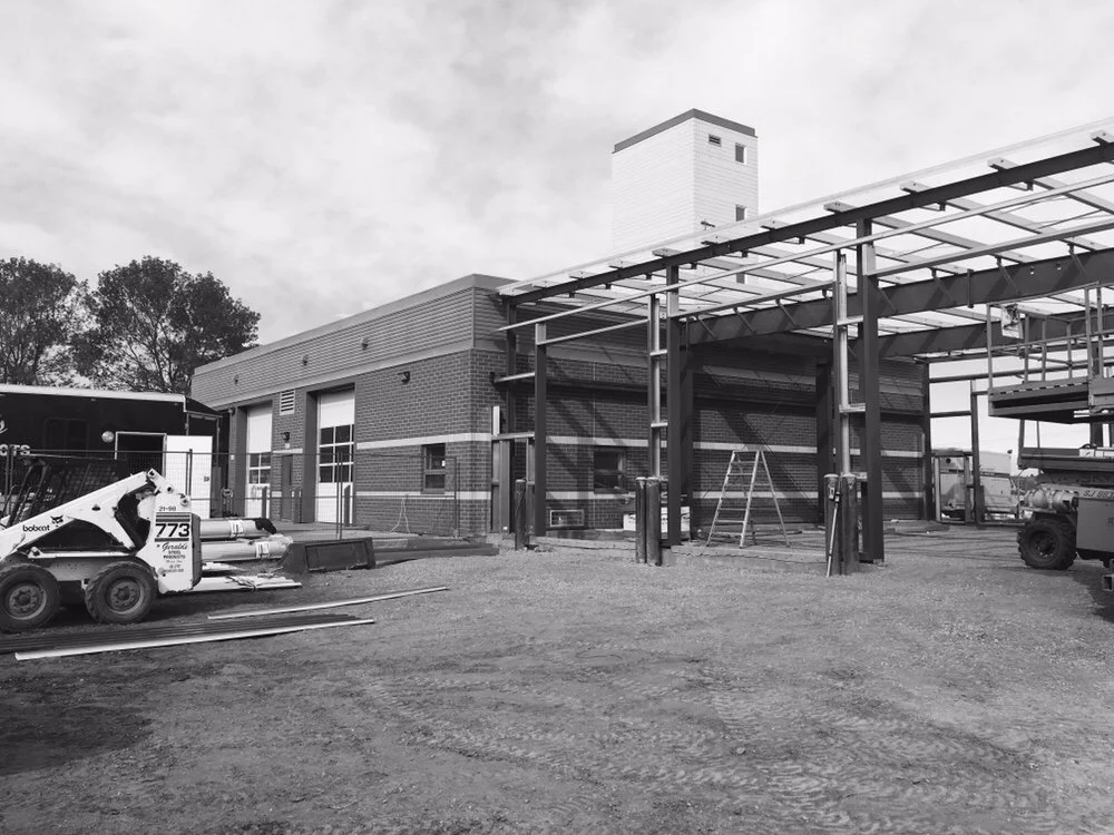 Construction site with a partially built structure featuring steel framing and a brick building in the background, construction equipment and materials present.