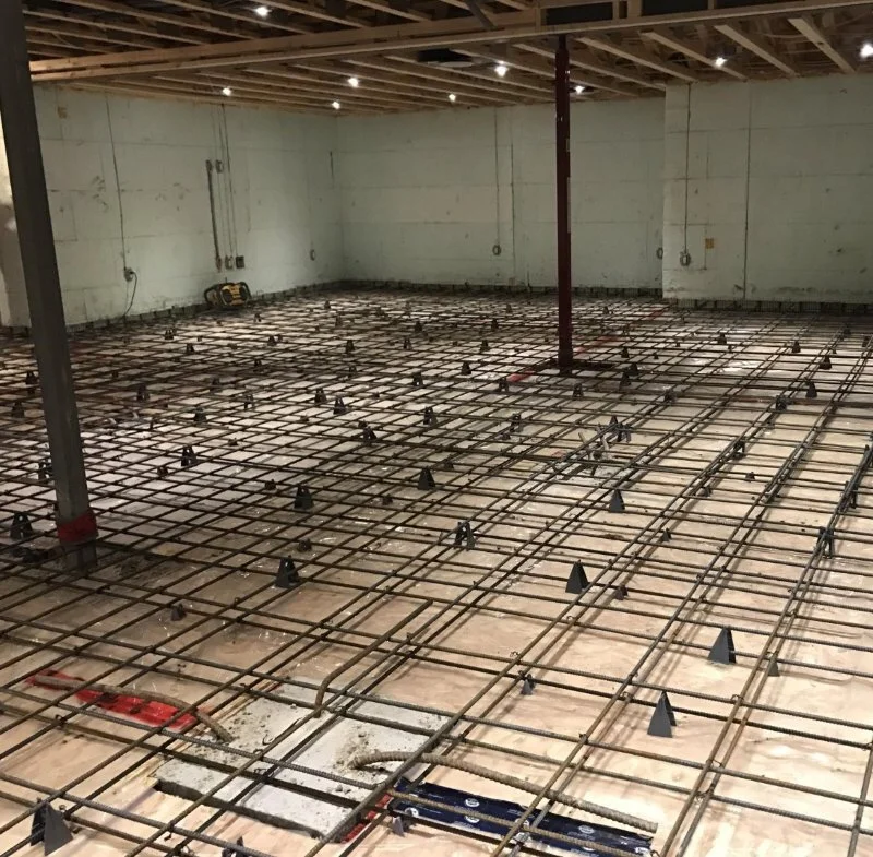 Interior of a building under construction with metal rebar grid on the floor, wooden ceiling, and unfinished walls.