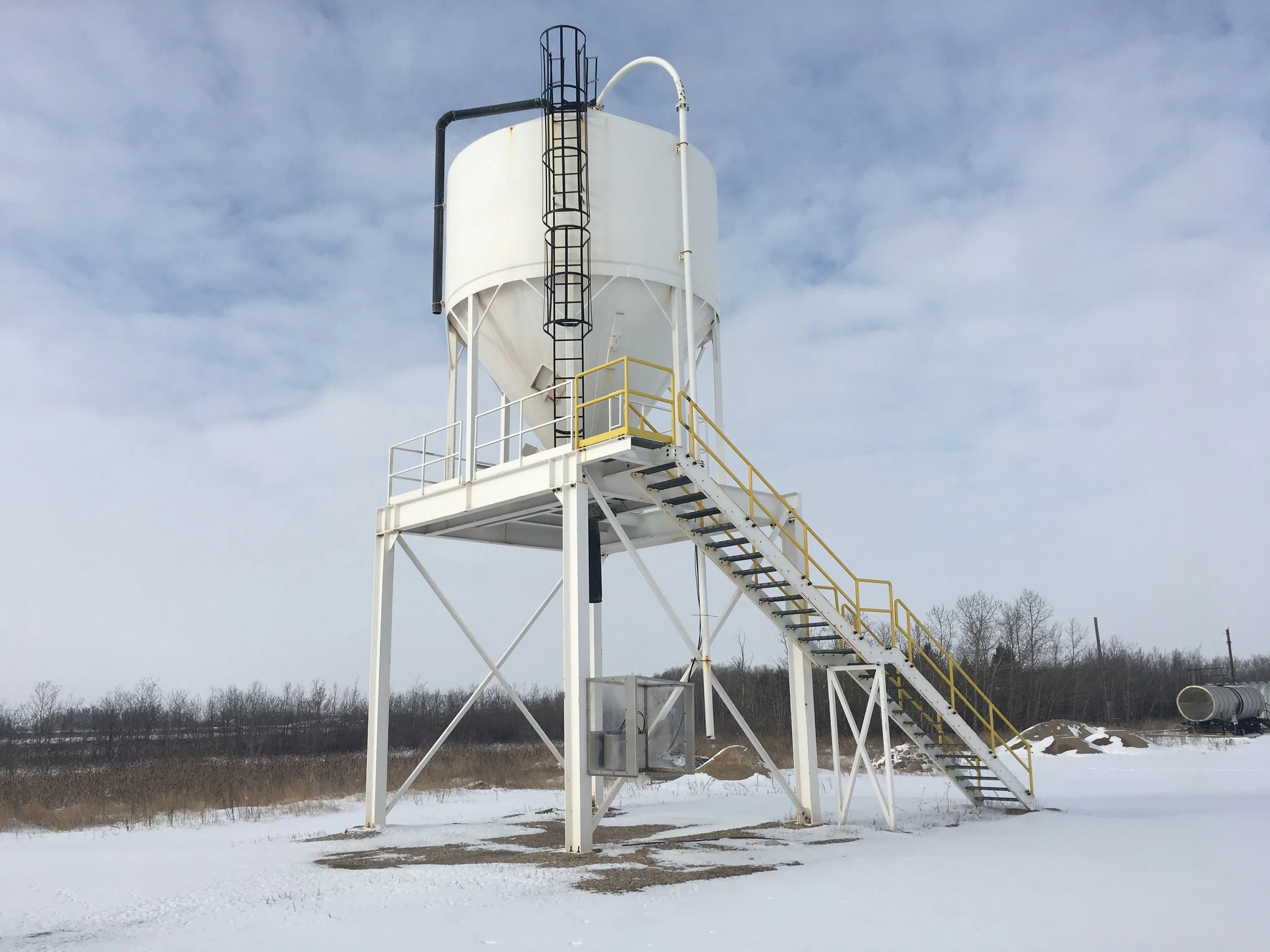 A tall white industrial silo with a black ladder and a staircase with yellow handrails, set against a cloudy sky. Snow covers the ground, with some trees and a barrel in the background.
