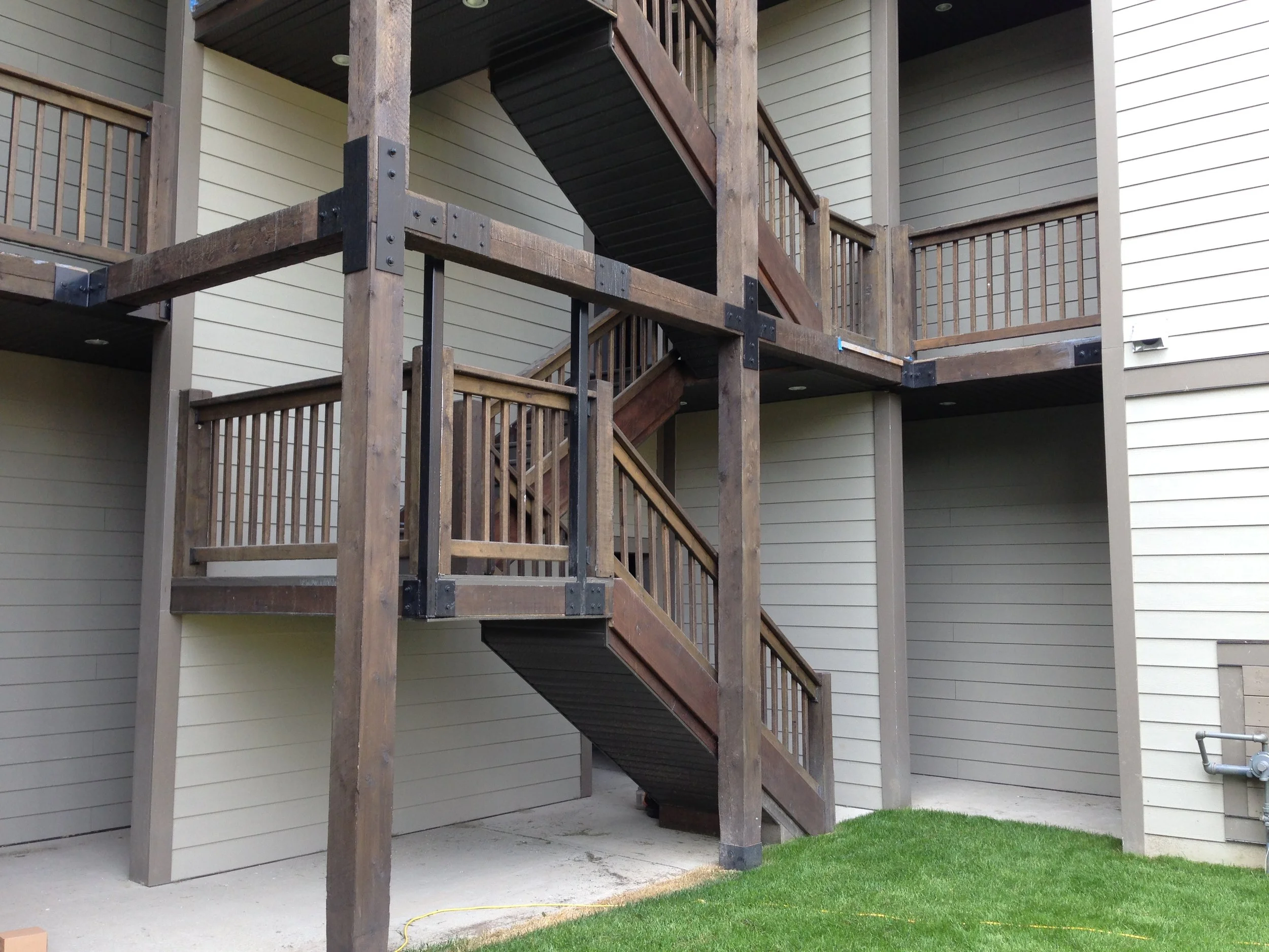 A multi-story wooden apartment building with an external staircase, beige horizontal siding, and a patch of green grass in the bottom right corner.