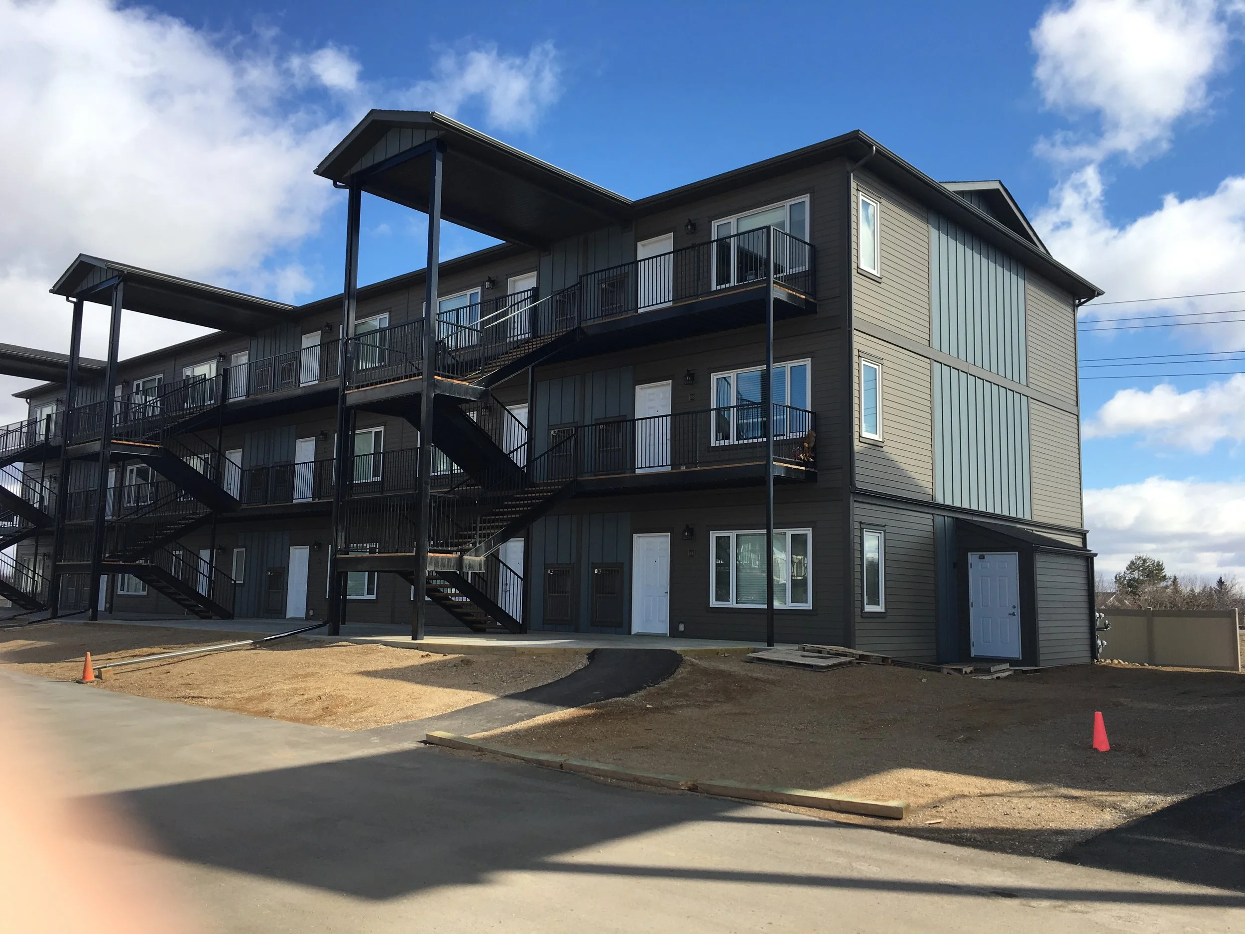 A multi-story residential building with exterior stairs, including black railings, under a blue sky with scattered clouds. The ground area is partially landscaped with dirt and some construction cones.