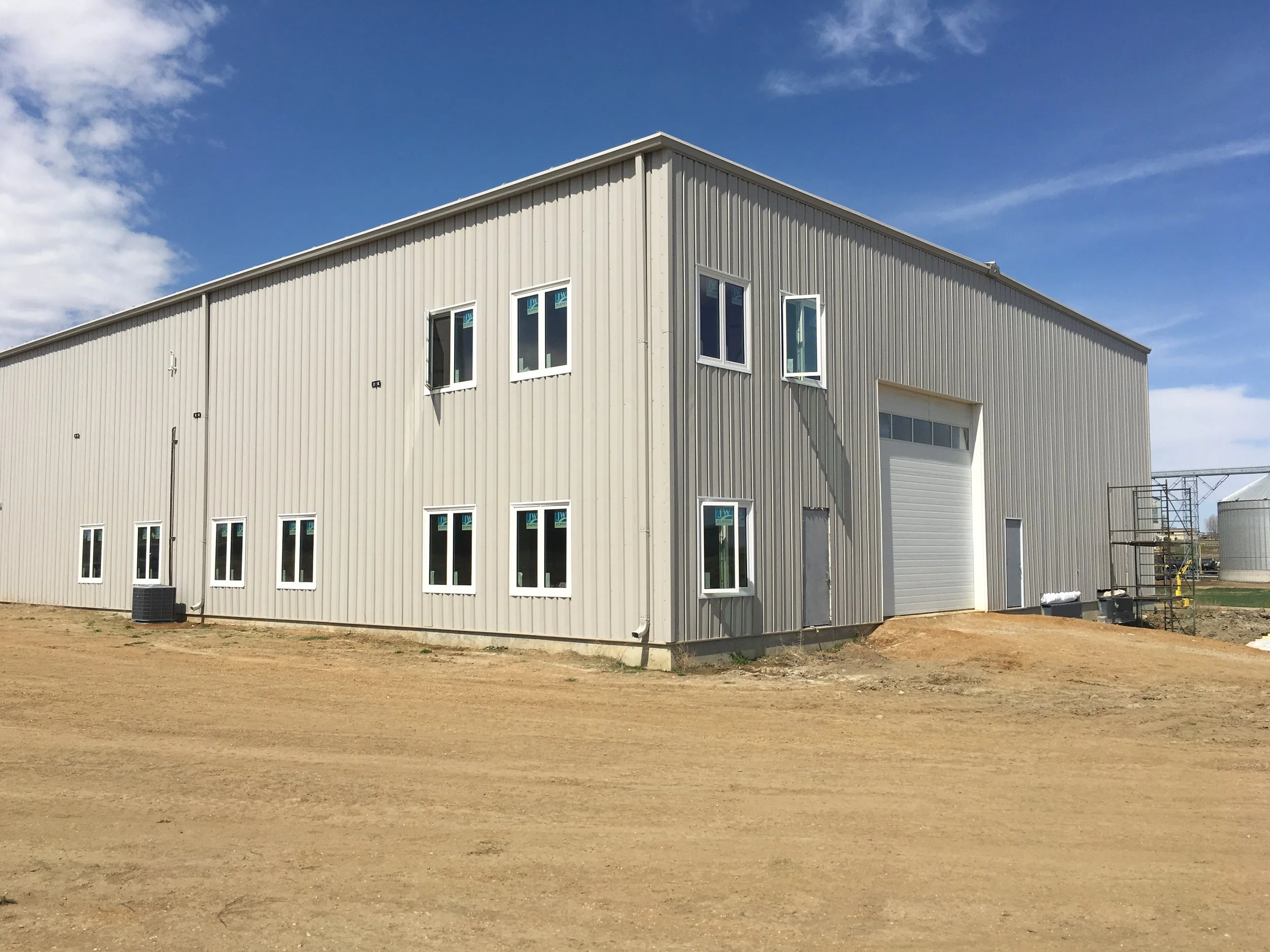 A large beige industrial building with multiple windows and a garage door set on a dirt lot under a partly cloudy blue sky.