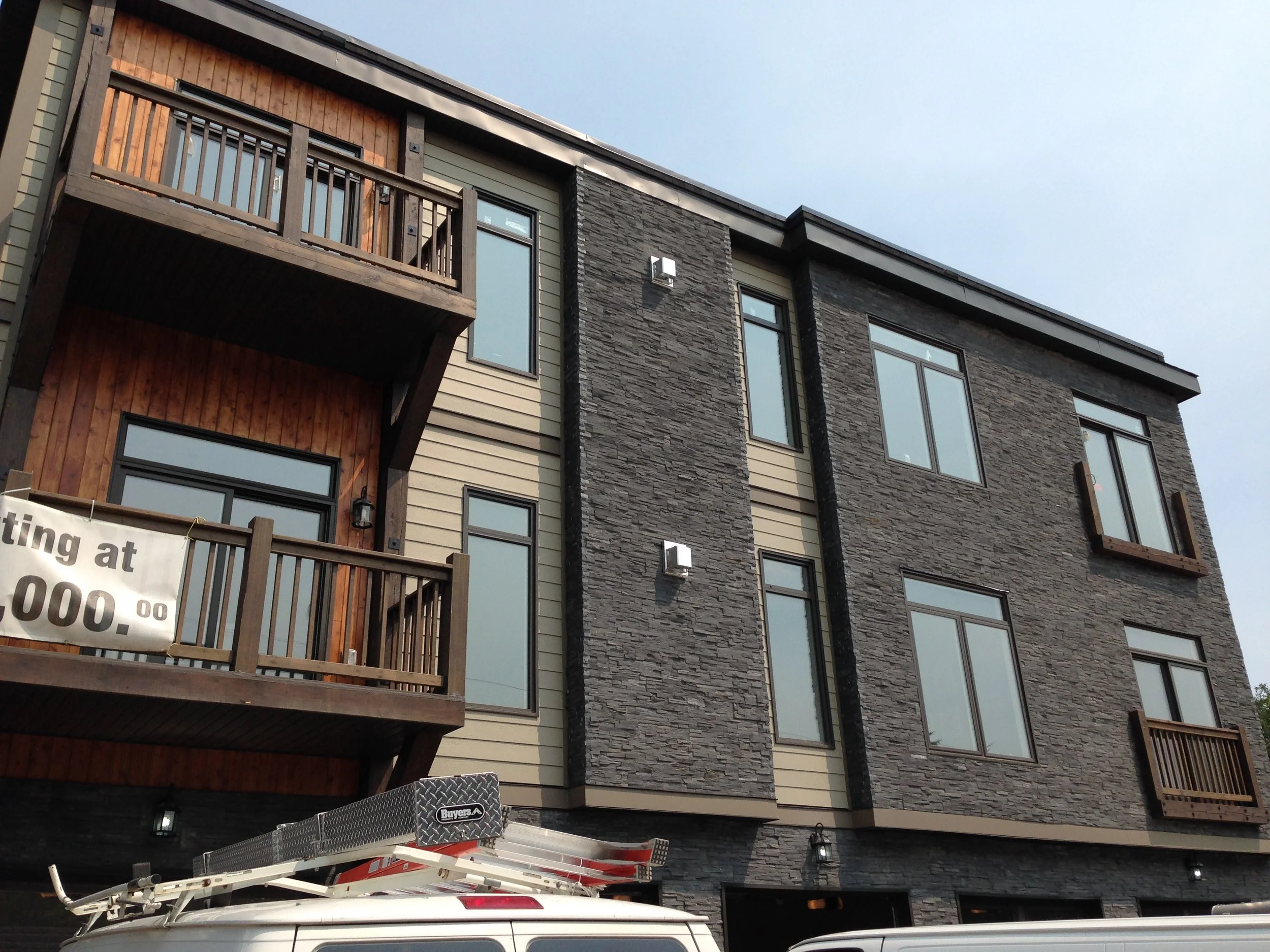 The image shows the exterior of a modern multi-story building with a combination of different materials on the facade, including wood, stone, and siding. There are several windows, including large windows, and small balcony spaces with wooden railings. A utility vehicle with a ladder or equipment on top is parked in front of the building, and part of a sign is visible in the lower left corner.