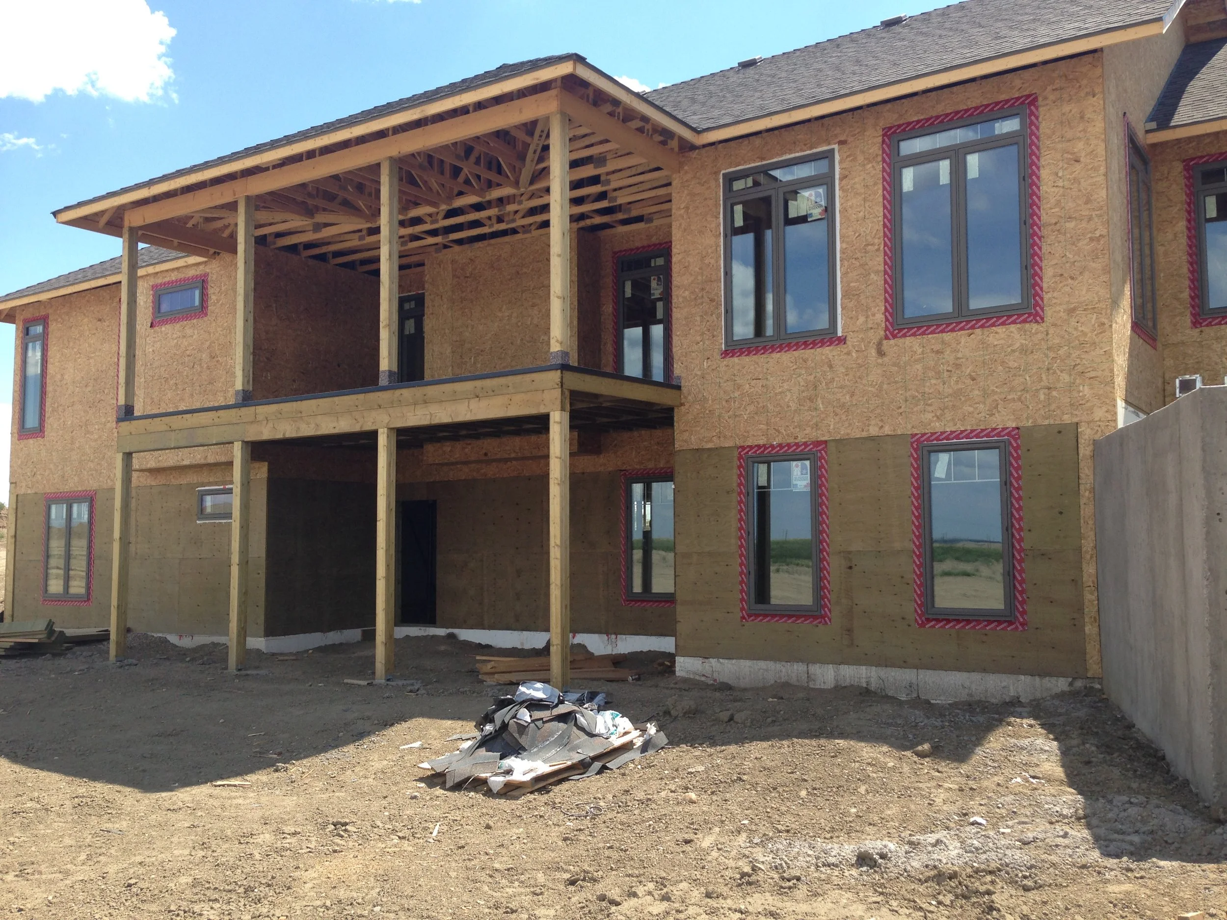 A house under construction with wooden framing, windows installed, and scaffolding, on a clear day with a bright blue sky.