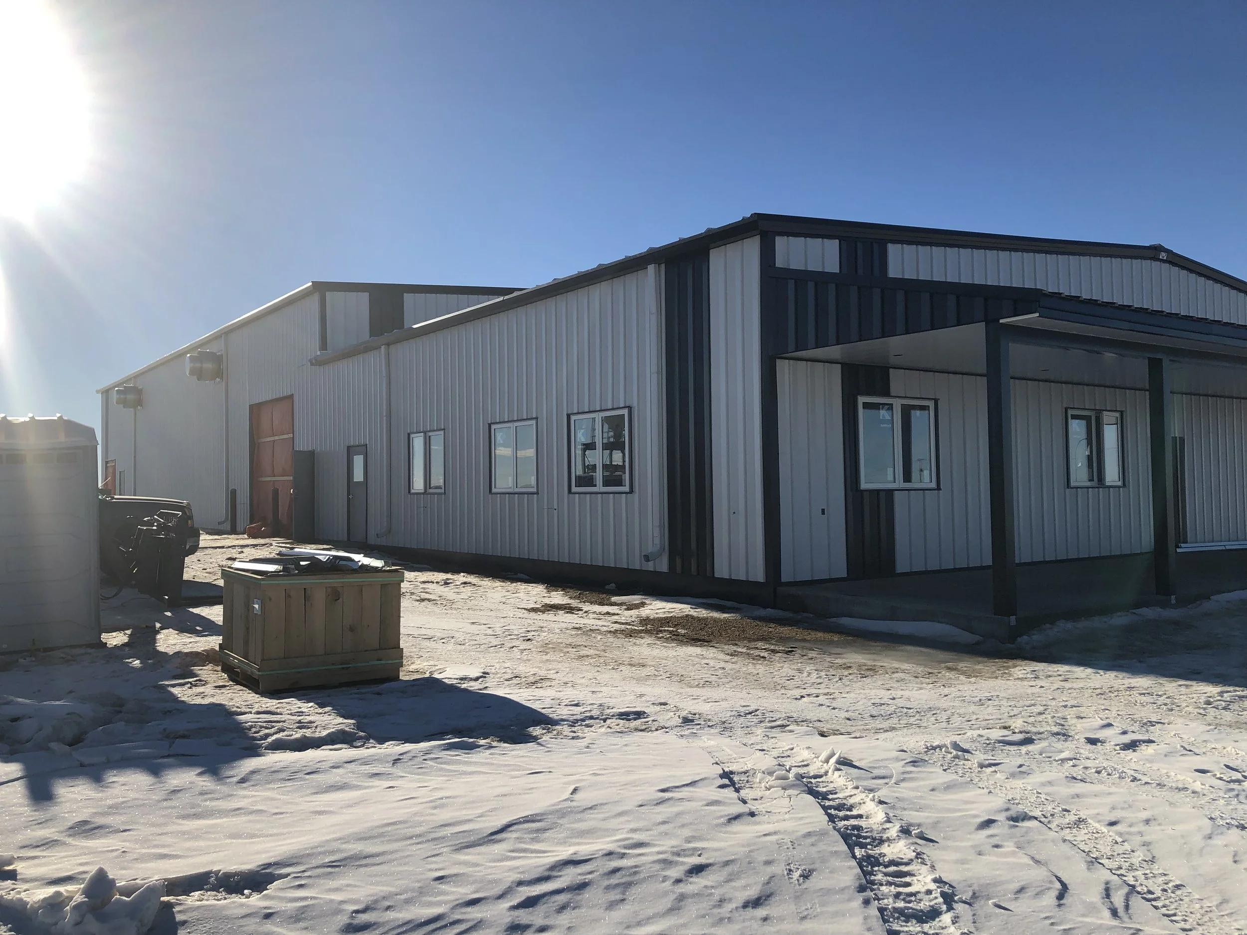 An industrial building with metal siding, several windows, and a covered entrance, located outdoors on a snowy ground under a clear blue sky with sunlight.
